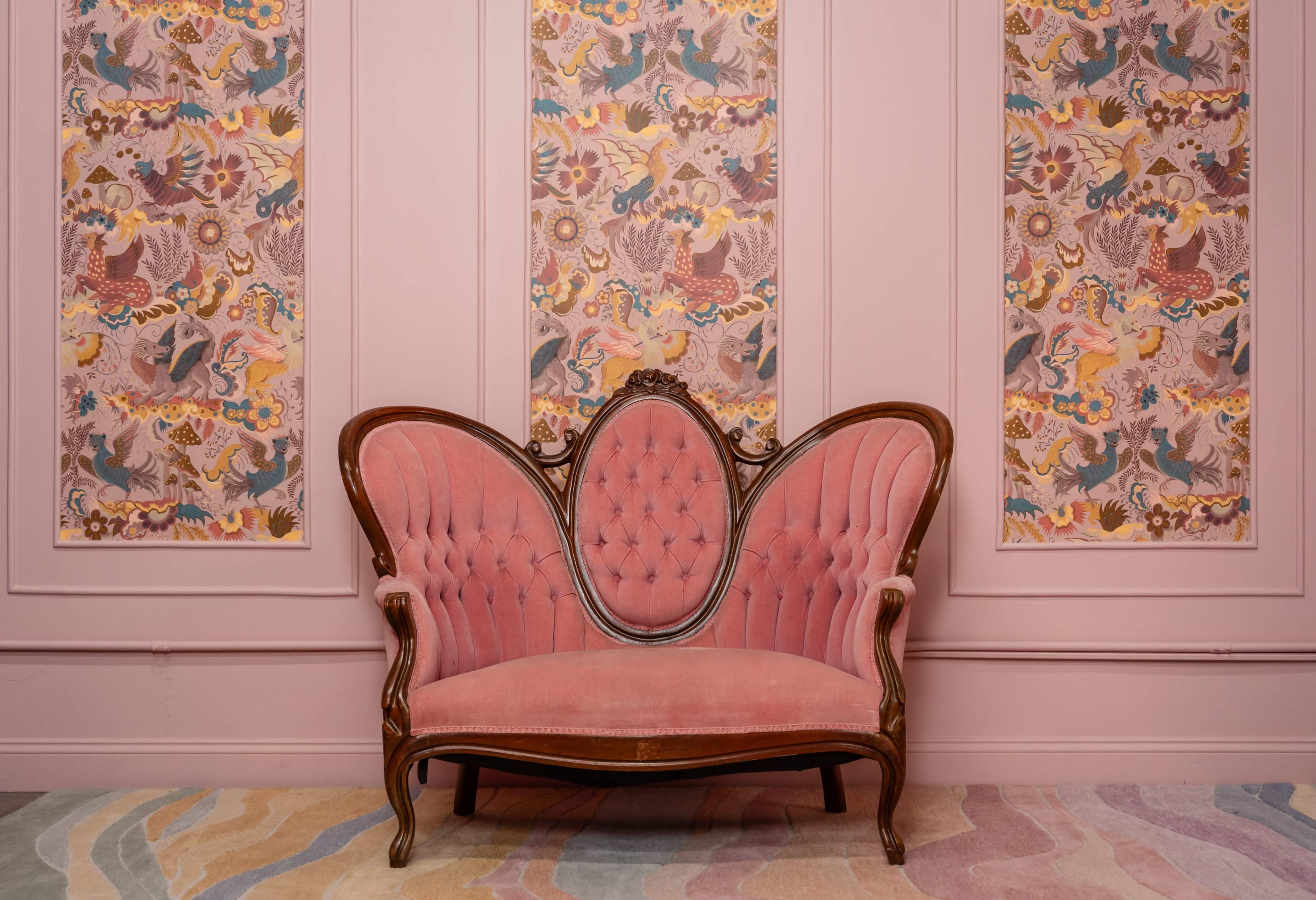 The image features an ornate pink upholstered sofa set against a wall with decorative floral and animal patterned wallpaper.