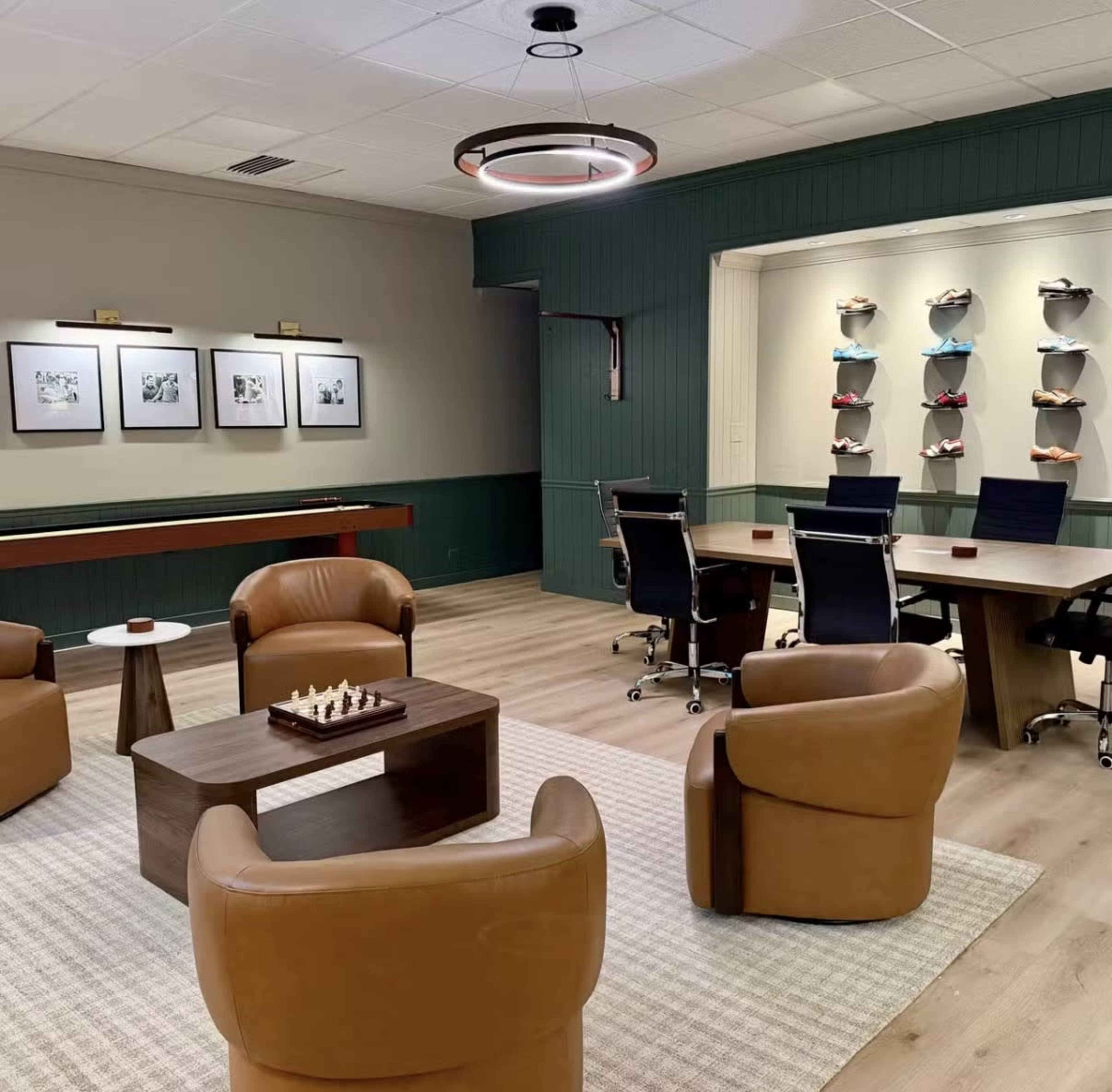 The image shows a modern office lounge area featuring four brown leather chairs, a wooden coffee table with a chess set, and a conference room in the background with a wall display of colorful shoes.