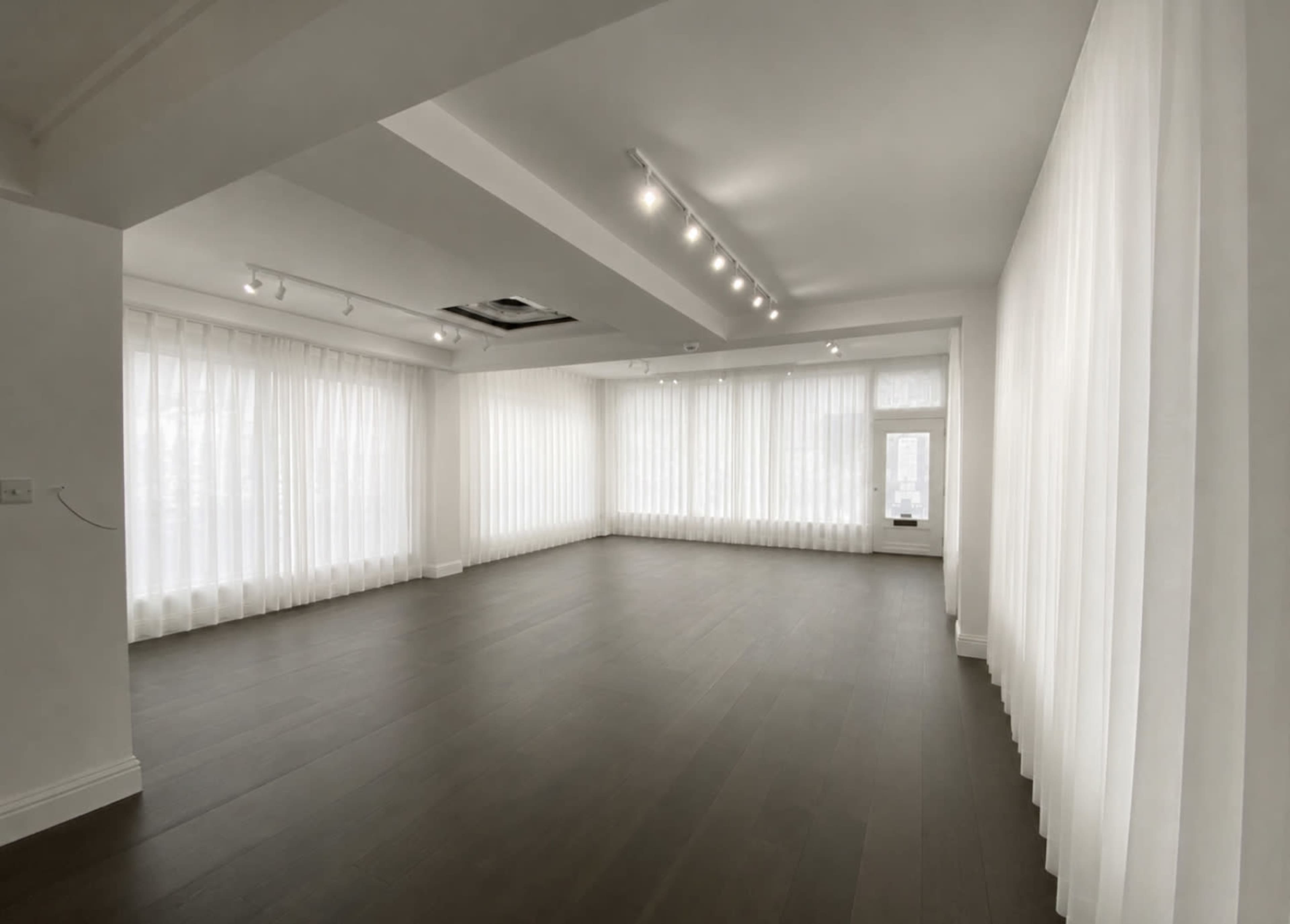 The image shows a spacious room with light-colored walls and large windows draped with sheer white curtains, featuring a dark wooden floor.