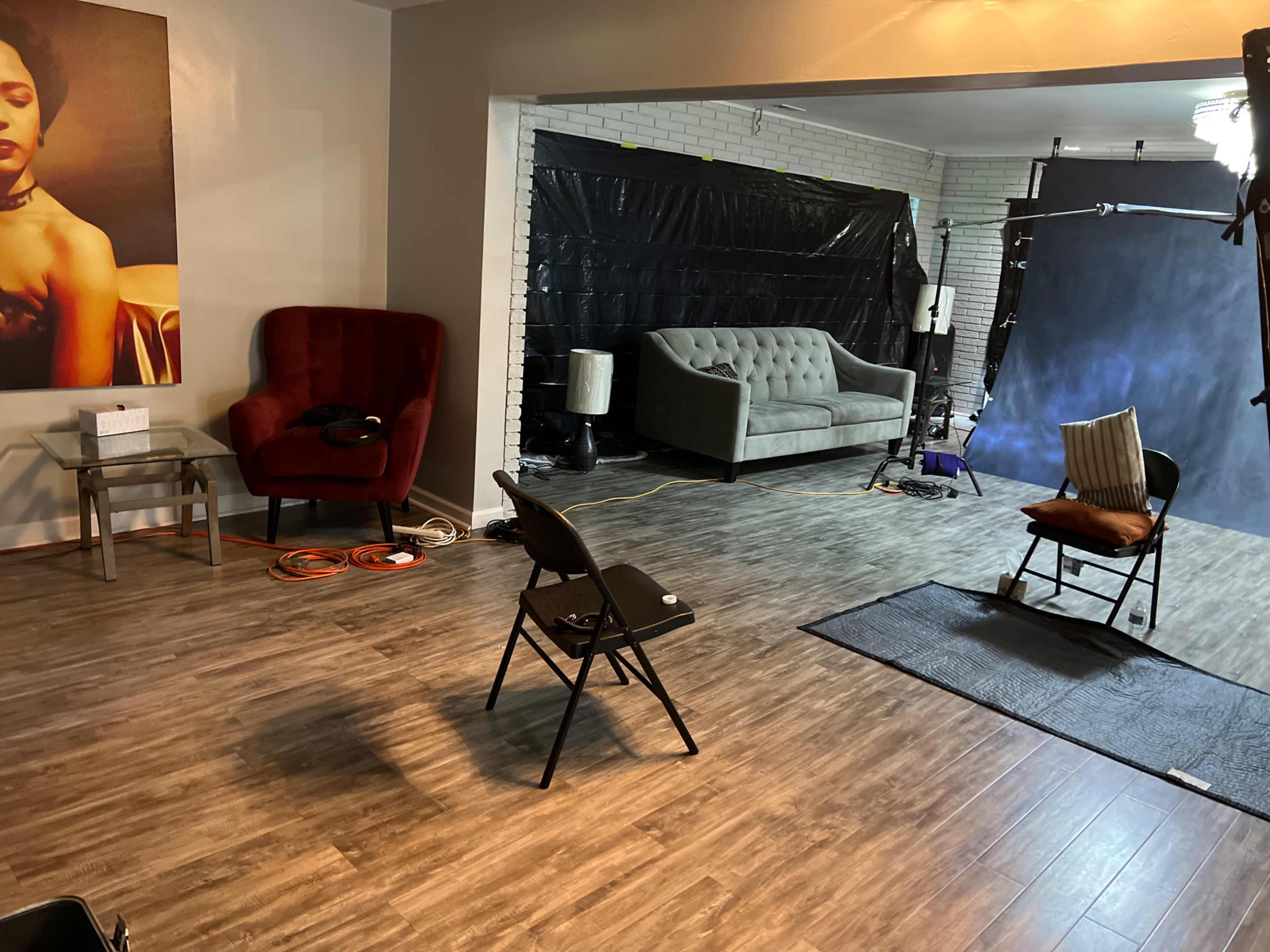 The image shows a professional studio space with a red velvet armchair, a gray couch, and various equipment set up for a photoshoot.