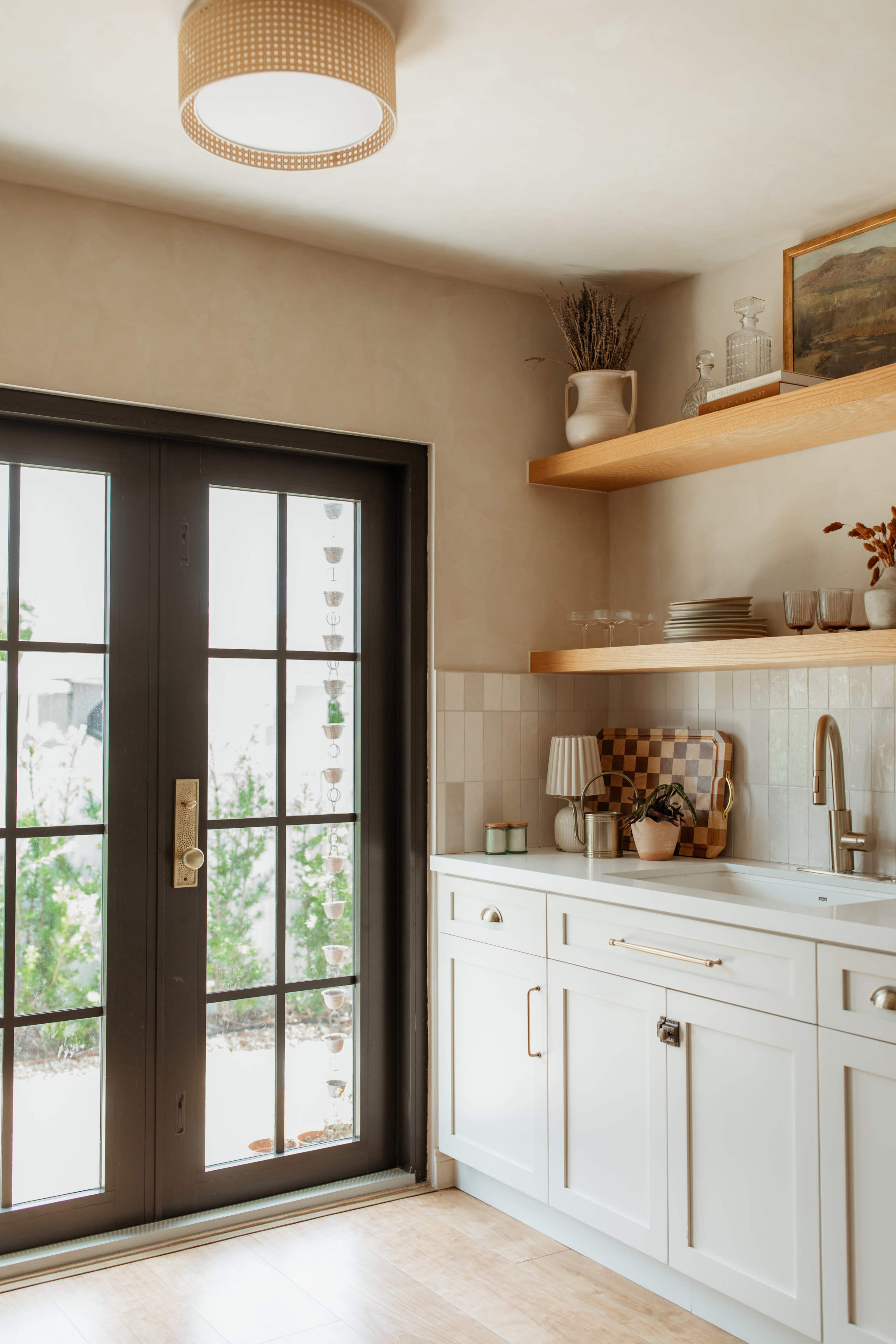 The image shows a well-lit kitchen featuring a black door with glass panes, white cabinetry, a wooden shelf with dishes, and a countertop with a sink.
