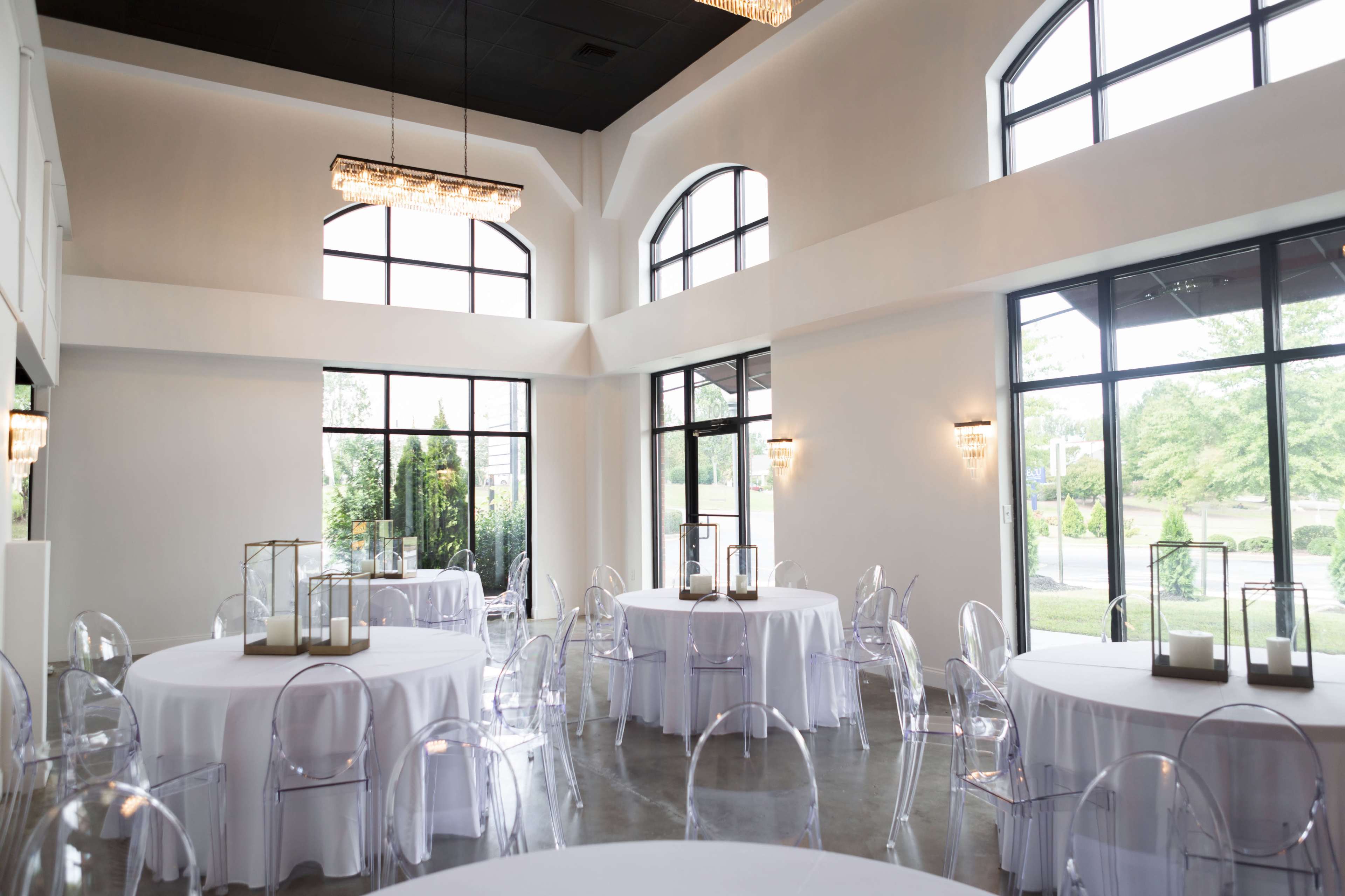 A bright event space with several round tables set, clear chairs, and large windows allowing natural light.