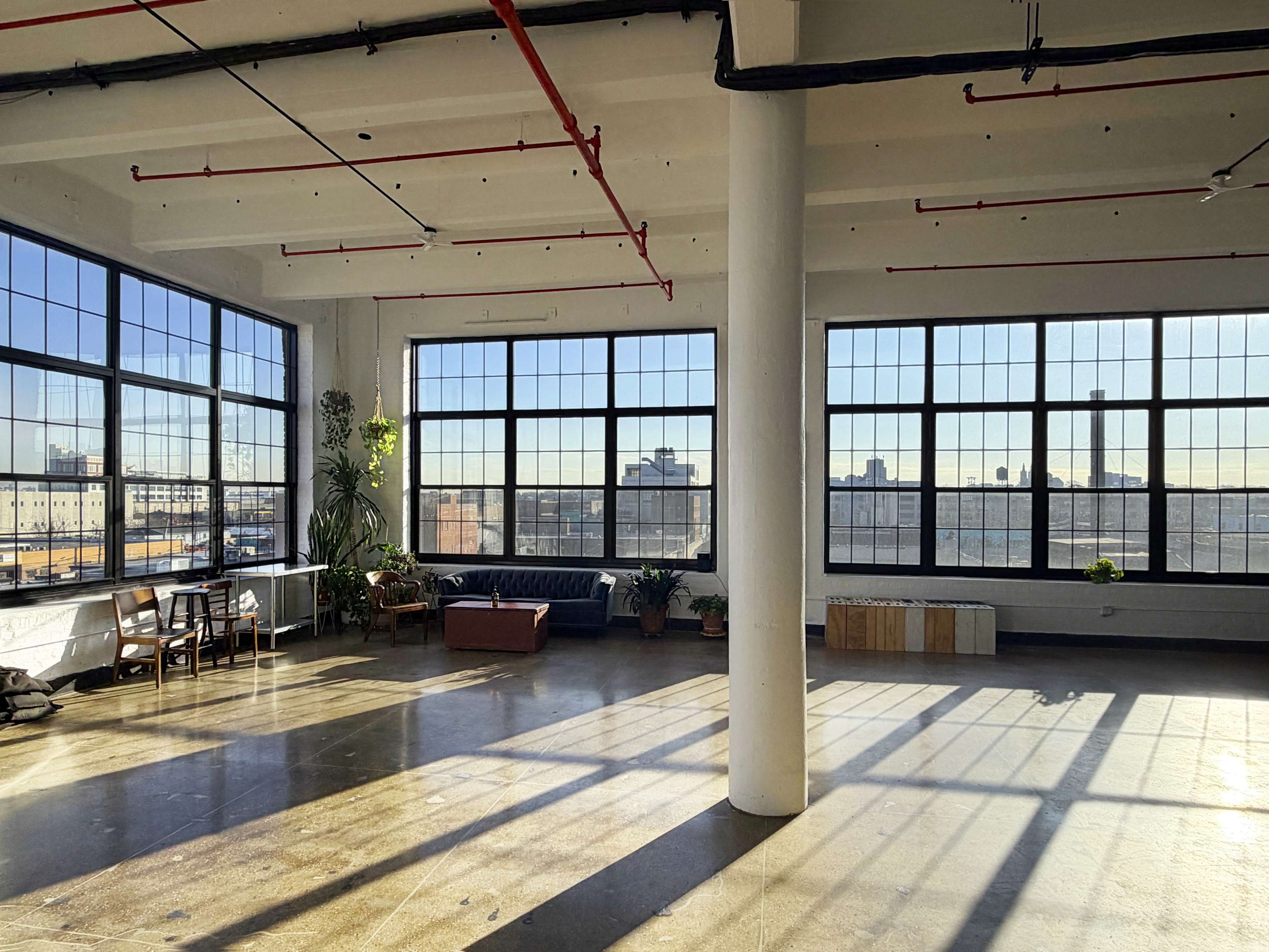 The image shows a spacious, well-lit industrial loft with large windows, hardwood floors, and potted plants scattered around.