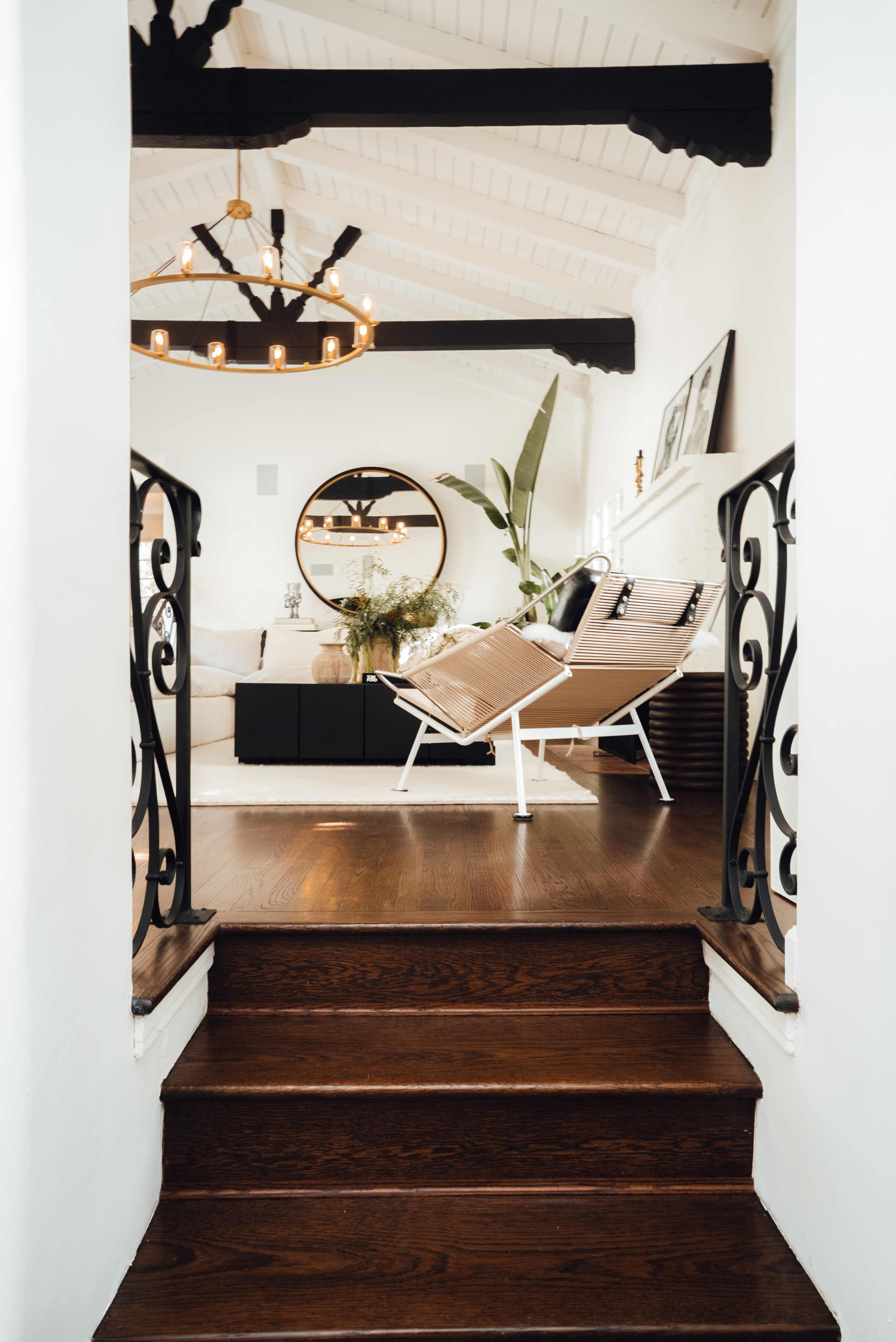 A set of wooden stairs leads down to a bright living room featuring a round mirror, a black cabinet, and two angled lounge chairs.