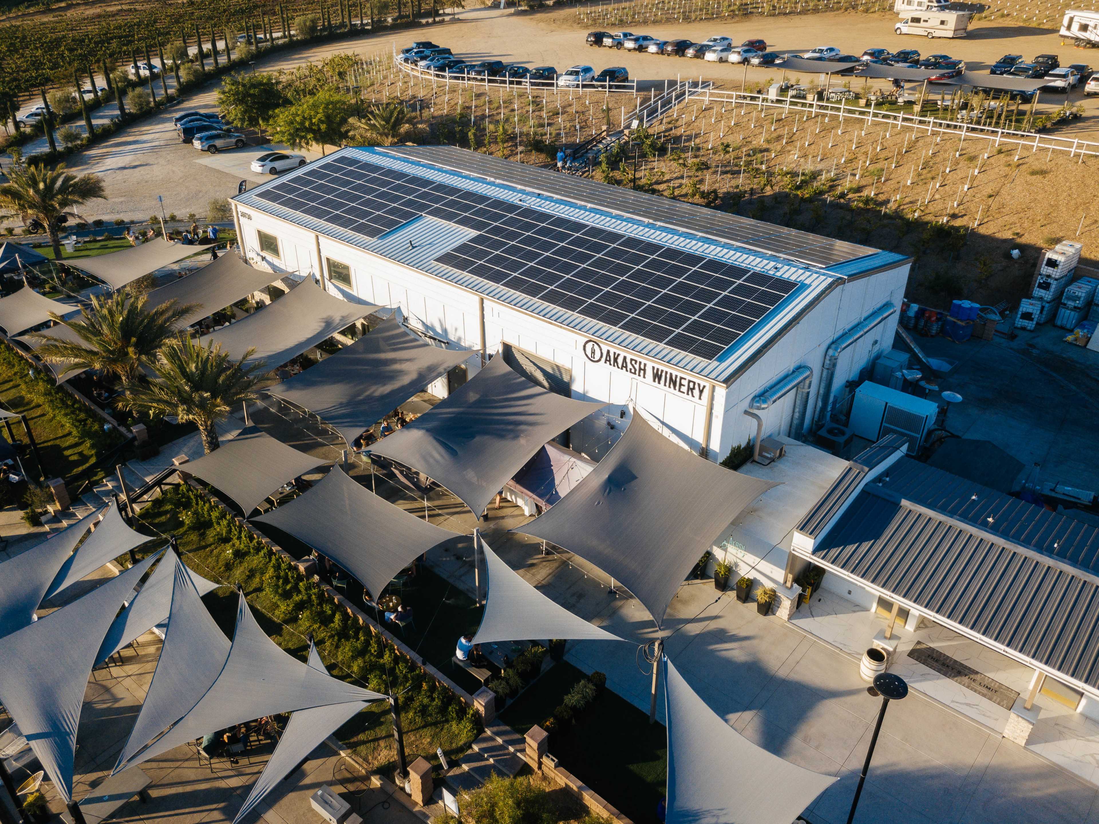 A winery building with solar panels on the roof, surrounded by outdoor seating areas covered by large fabric canopies.