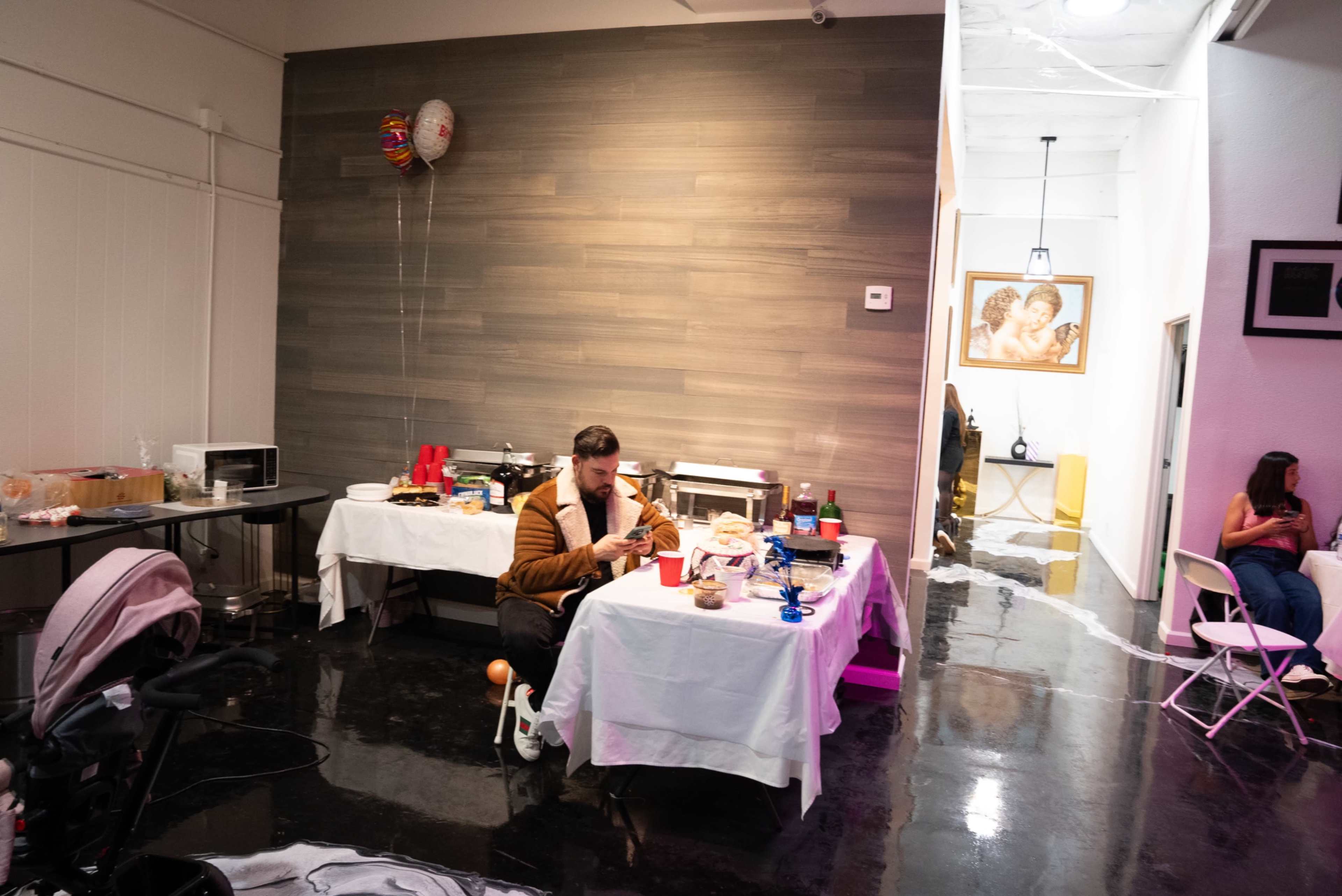 A man sits at a table in a dimly lit room filled with food, while another person relaxes nearby in a chair.