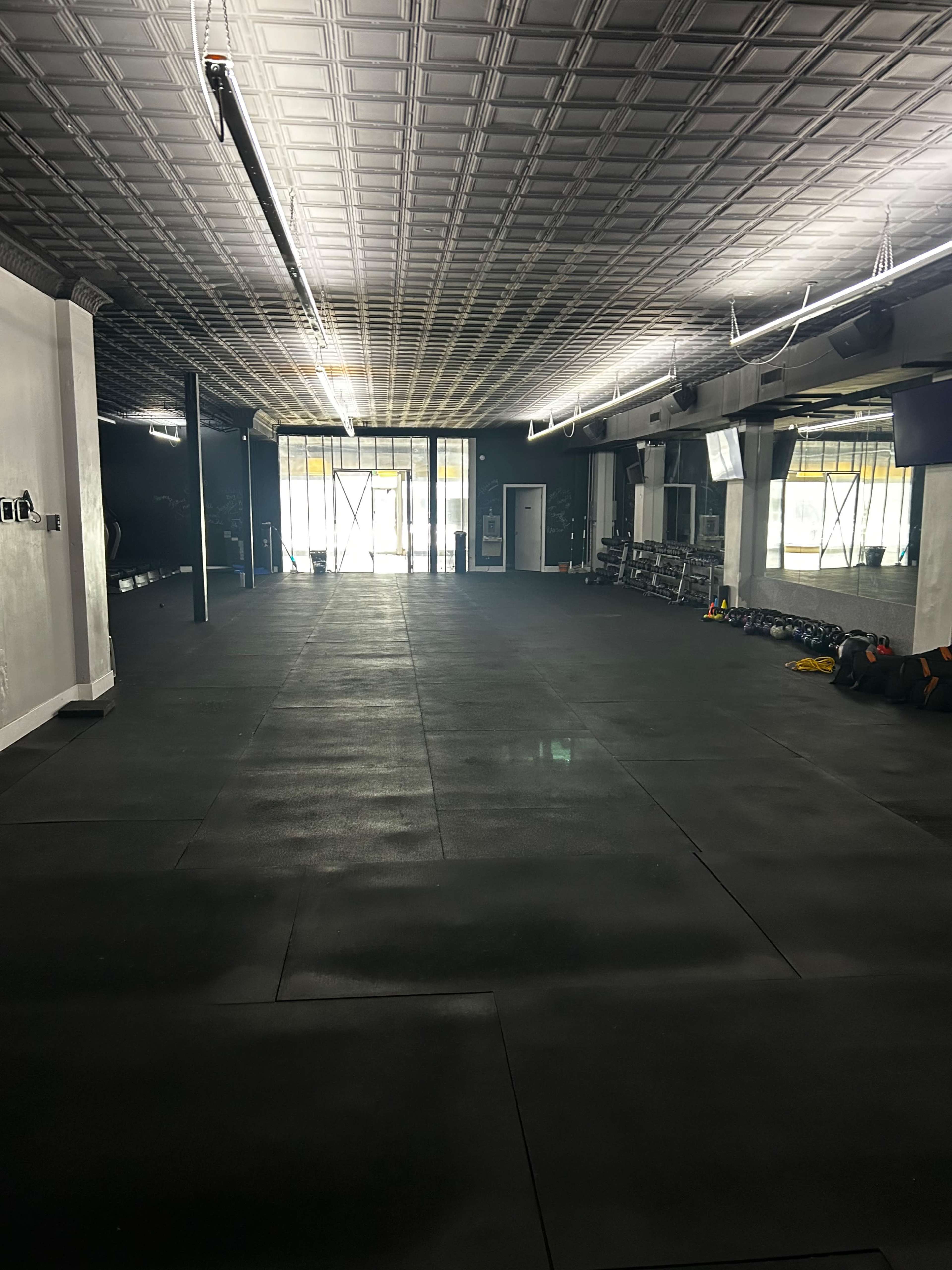 The image shows a spacious, empty gym with a black rubber floor and large windows allowing natural light to enter.