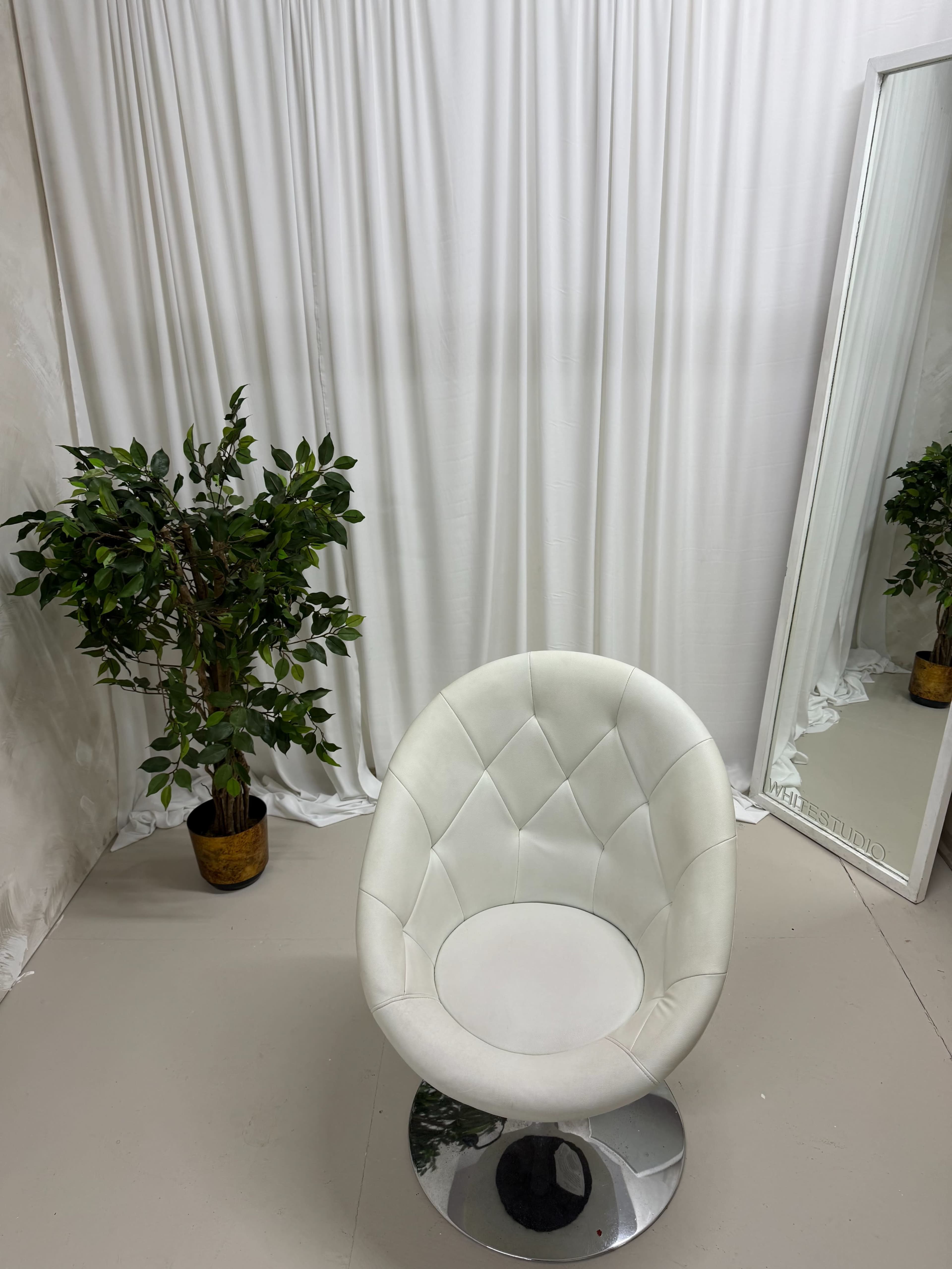 The image shows a white padded chair in front of a mirror, with a small potted plant beside it against a backdrop of draped white fabric.