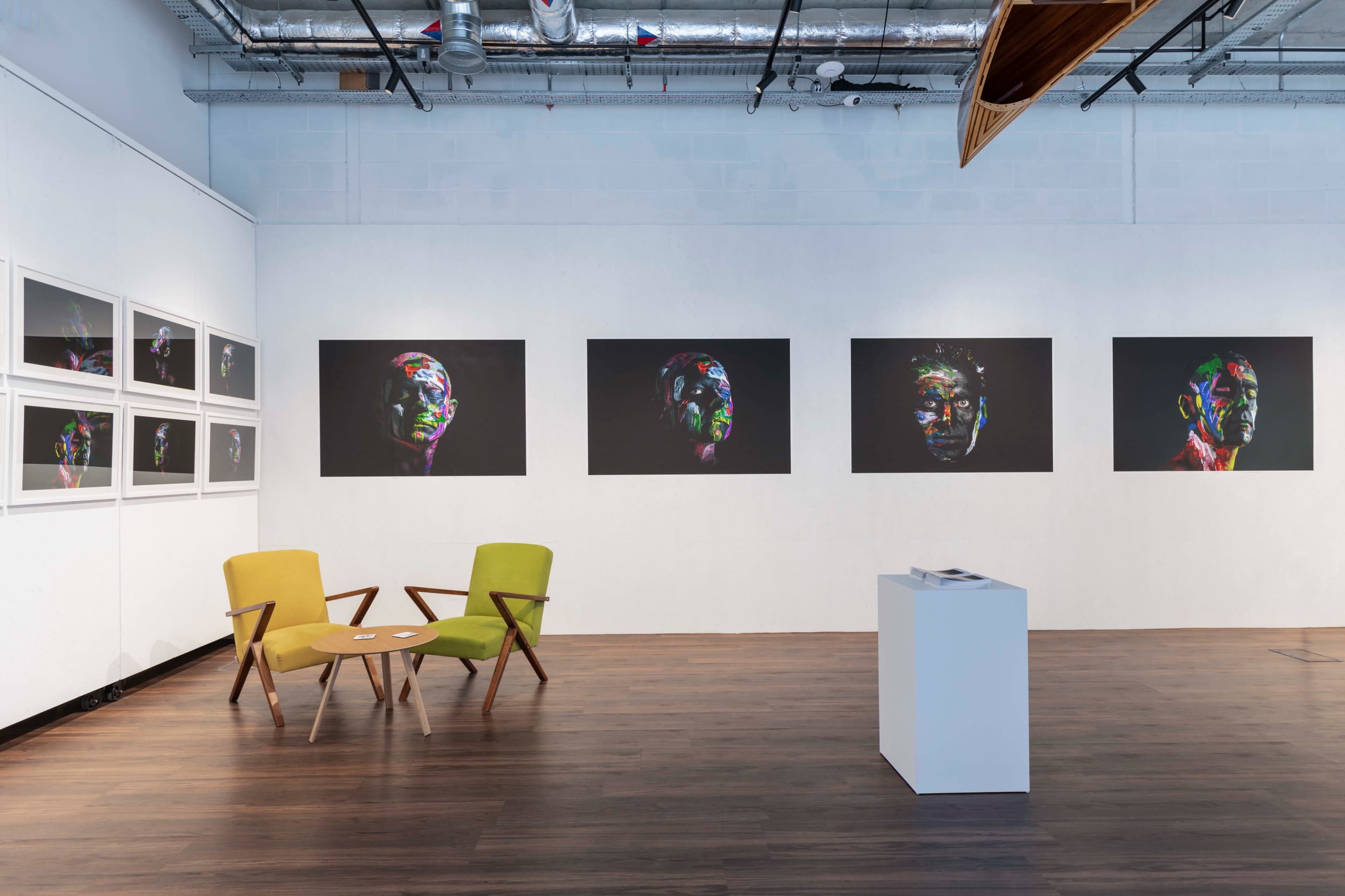 An art gallery featuring colorful portrait photographs on the walls, with a pair of modern chairs and a small white table in the foreground.