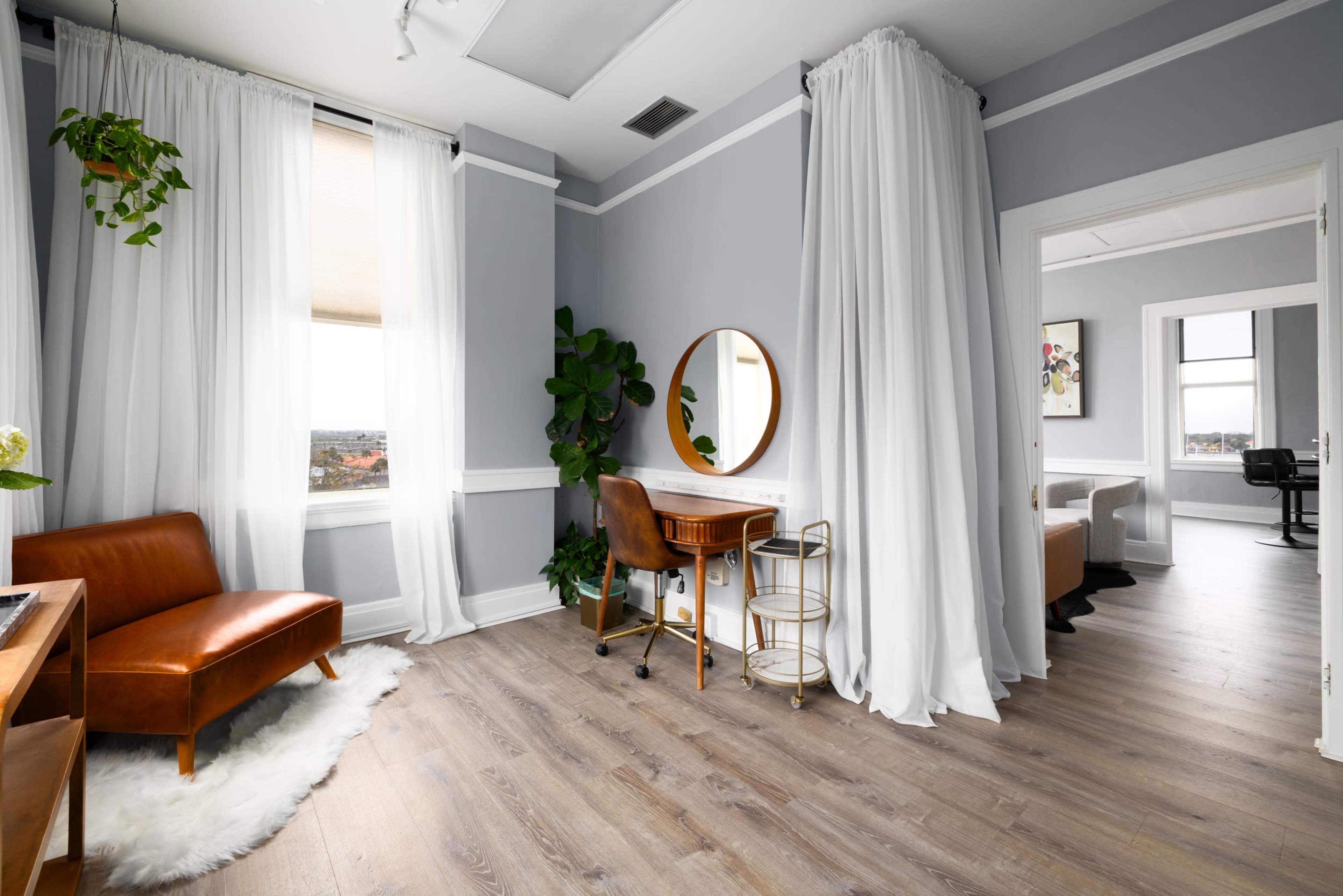 The image shows a modern interior of a light-filled room featuring a brown leather chair, a round mirror, a small metal cart, and white drapes, with wooden flooring and plants.