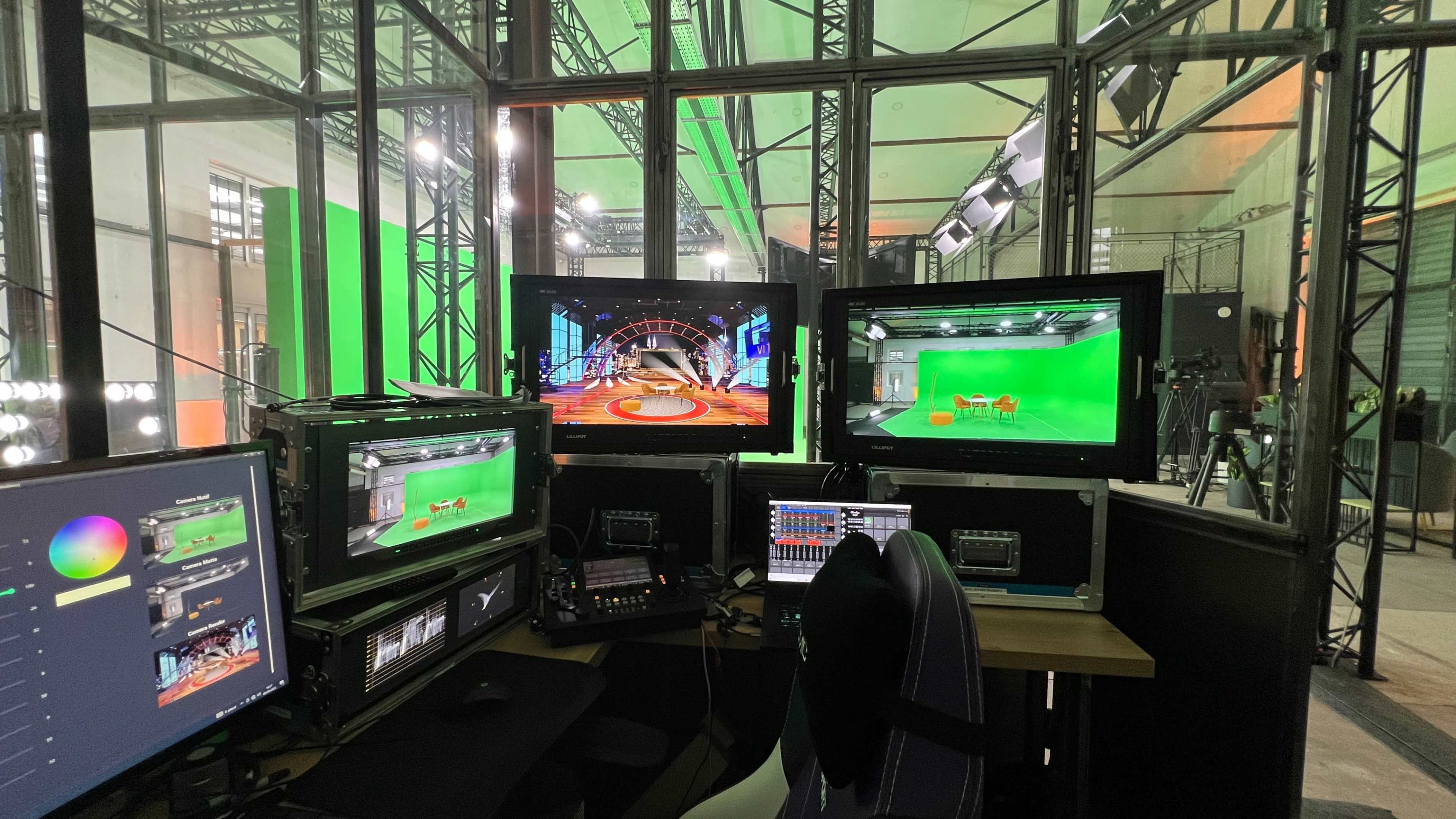 A control room with multiple computer monitors displaying virtual sets, audio equipment, and a green screen backdrop.