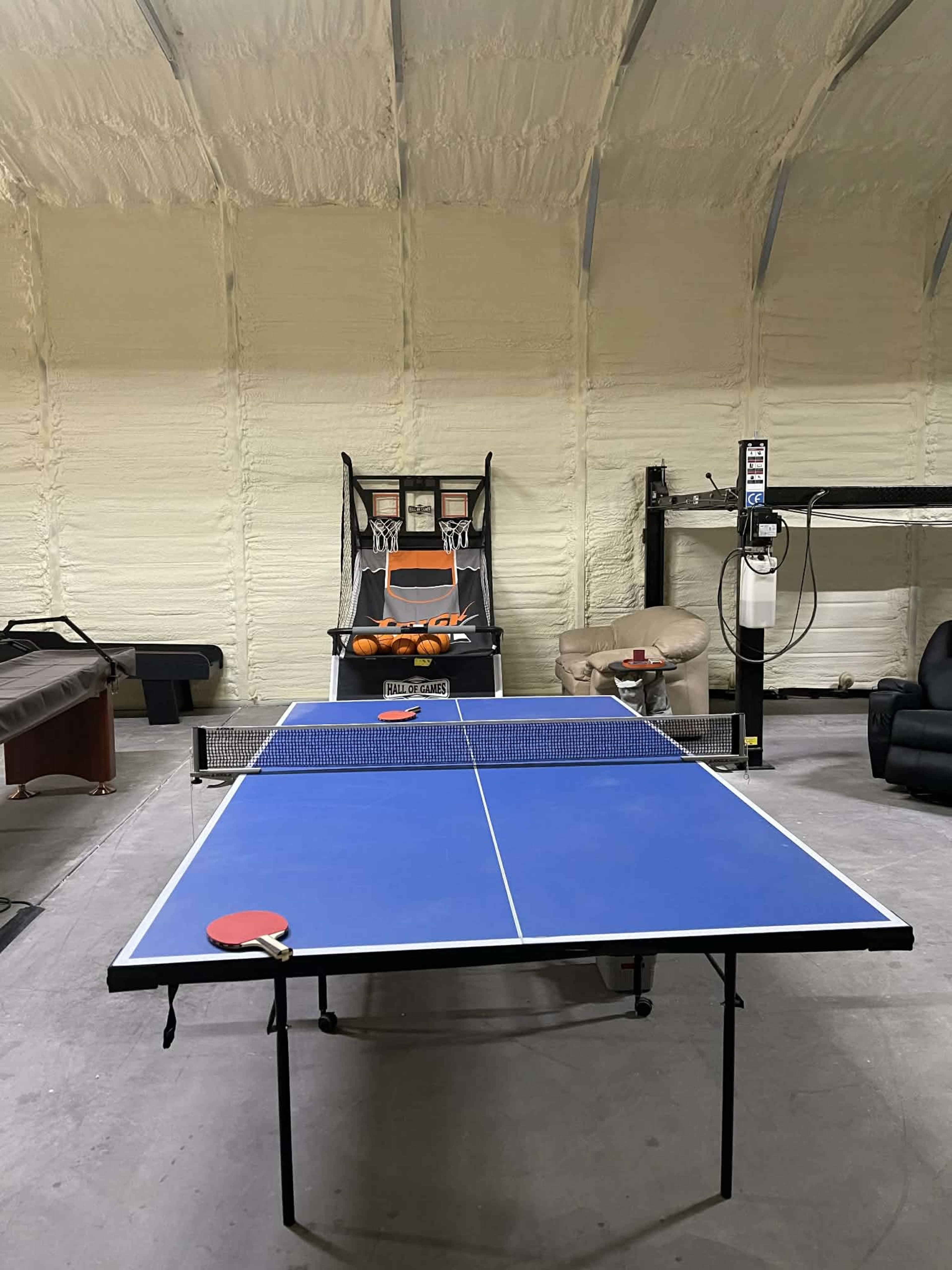 A blue ping pong table is set up in a spacious garage-like room, accompanied by various pieces of furniture including a couch and a table.