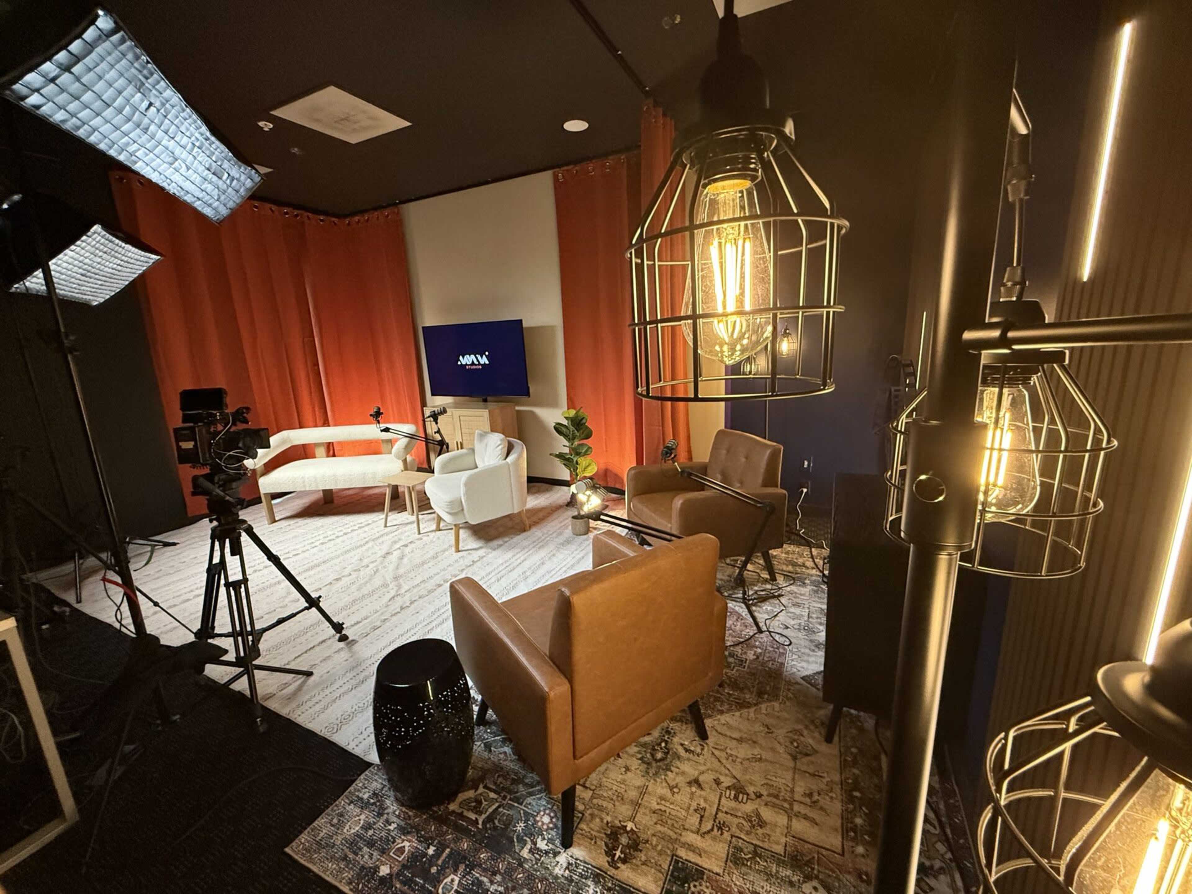 The image shows a modern studio setup with a camera, lights, and a television screen, featuring a comfortable seating arrangement with two chairs and a sofa surrounded by decorative elements.