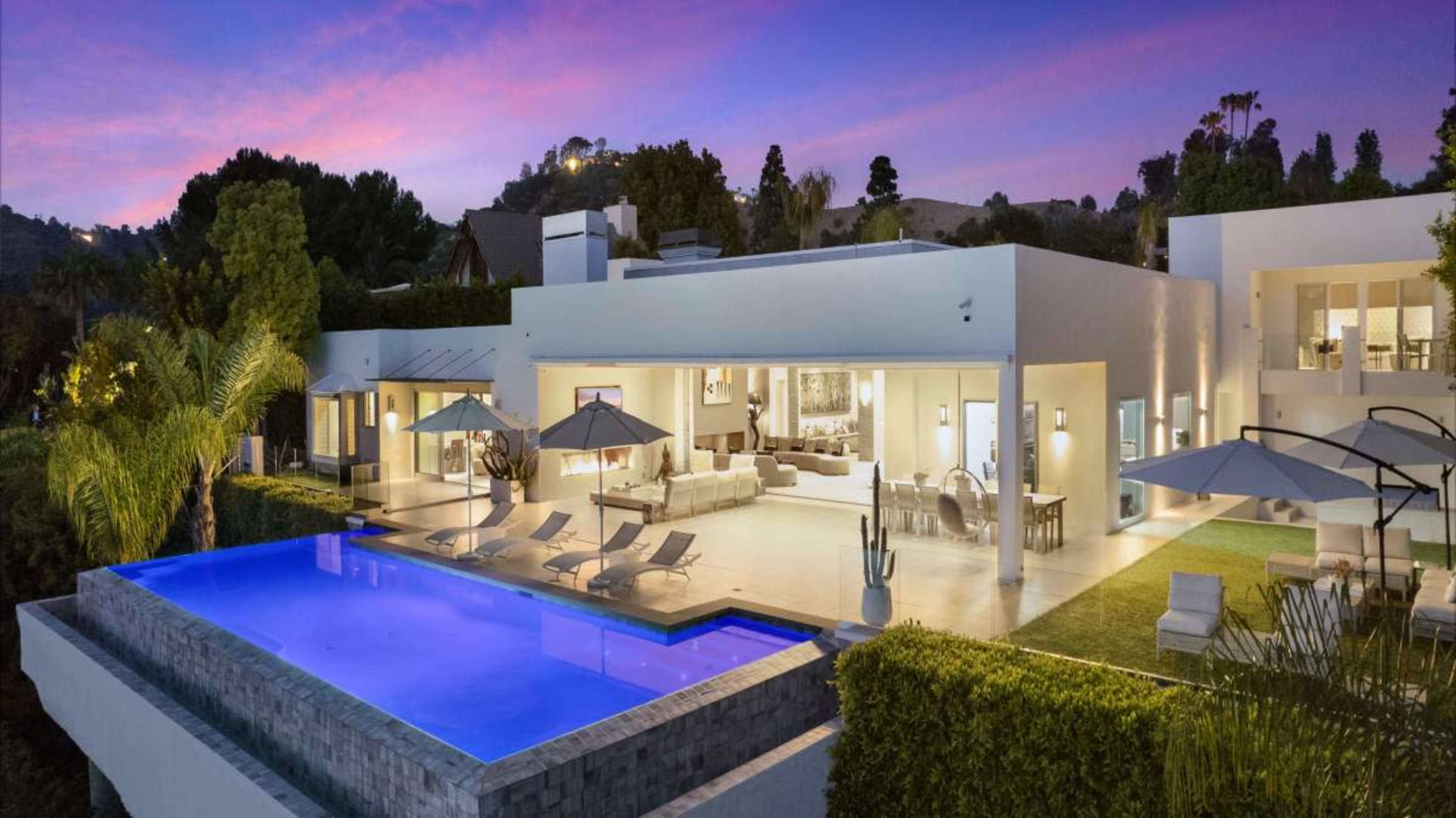 A modern villa features a spacious patio with a swimming pool and outdoor seating, surrounded by lush greenery against a twilight sky.
