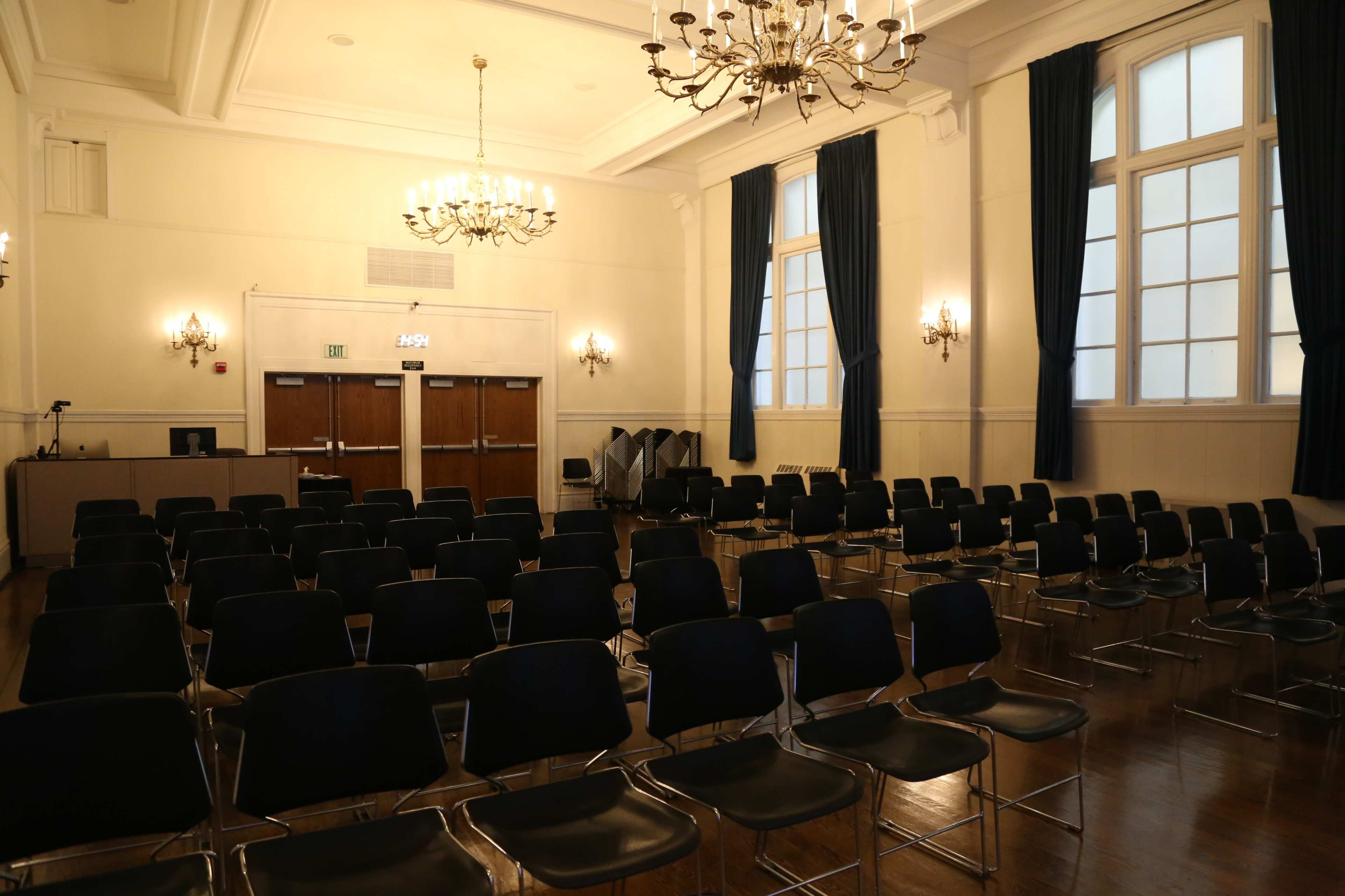 A spacious, well-lit room with rows of black chairs arranged in a semi-circle and large windows allowing natural light to enter.