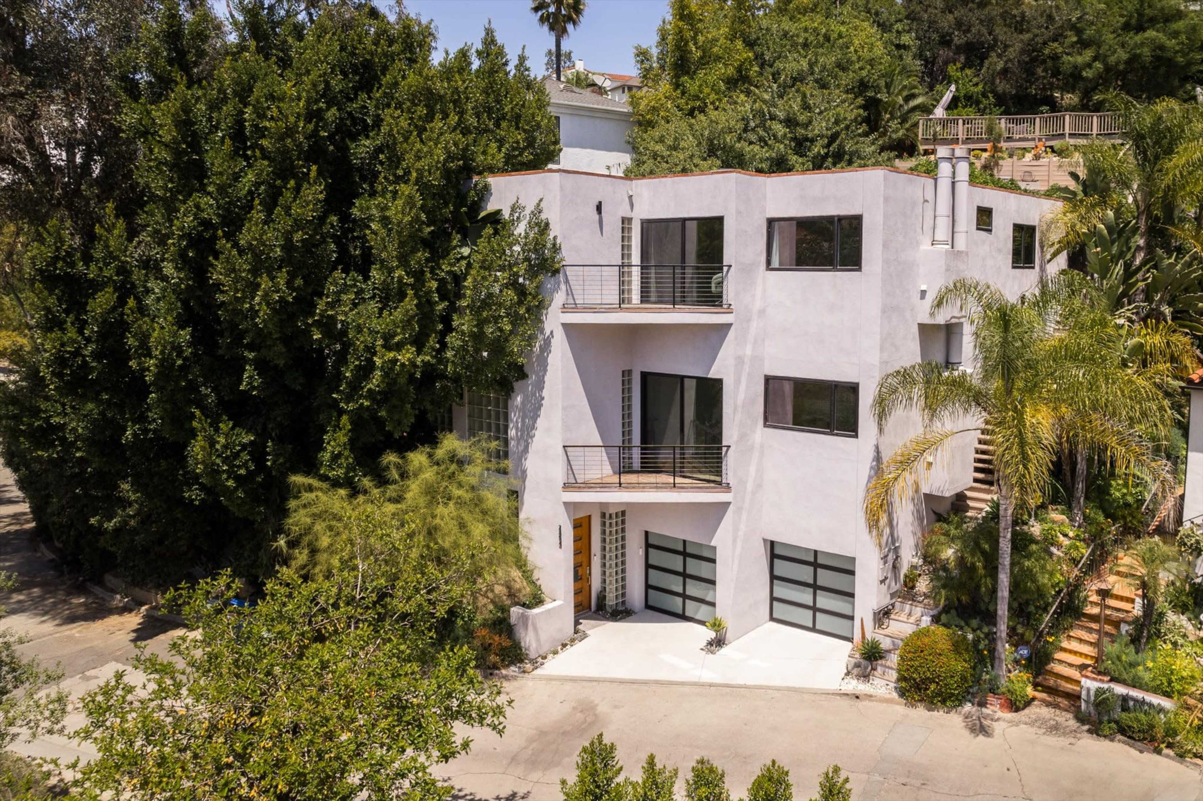 A modern, multi-level house is situated among lush greenery, featuring a combination of large windows and a driveway with two garage doors.