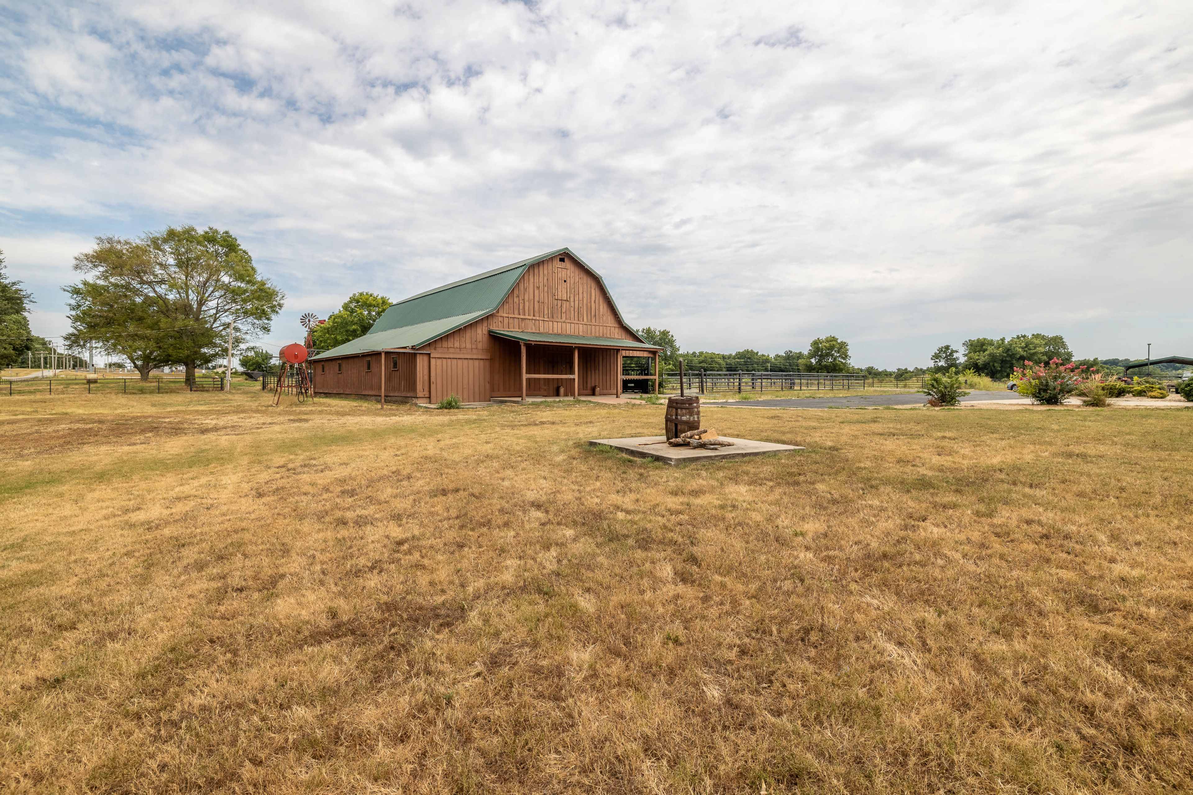 A large brown barn with a green roof stands in an open field with dry grass and a basketball hoop nearby.