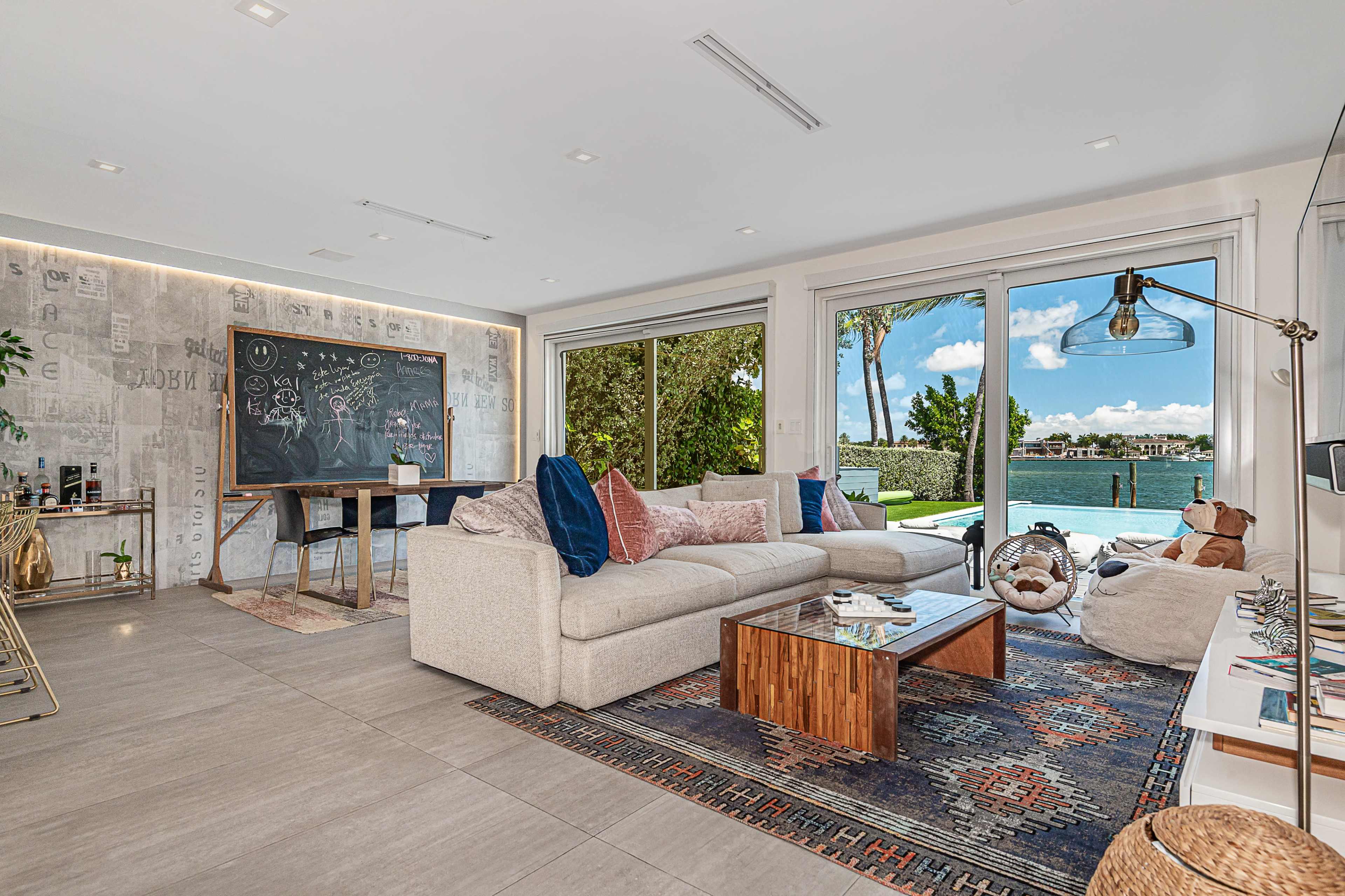A modern living room features a large sofa, a coffee table, a chalkboard, and large windows overlooking a waterfront view with palm trees.