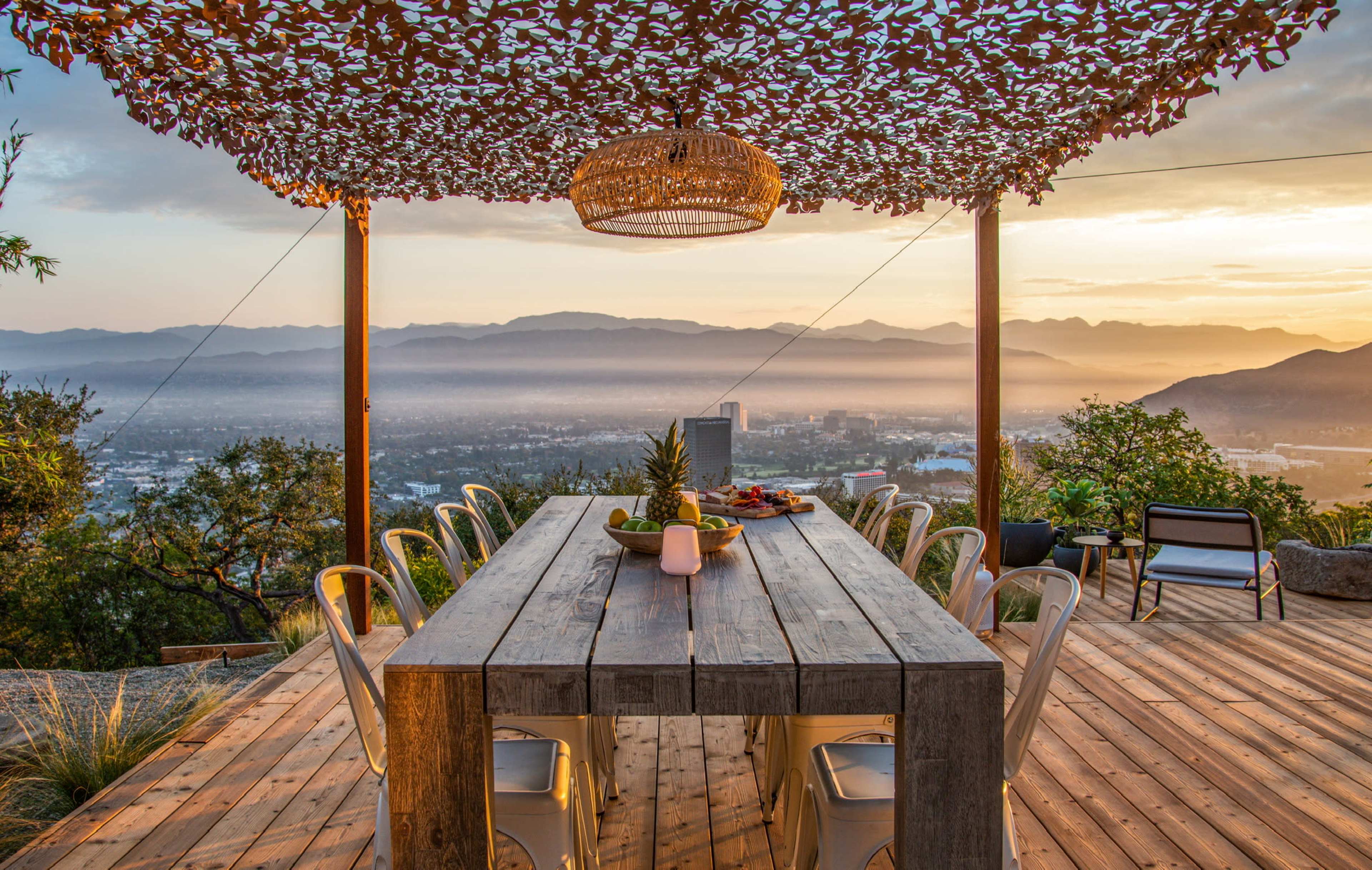An outdoor dining area with a wooden table and metal chairs is situated under a decorative canopy, overlooking a valley and mountains during sunset.