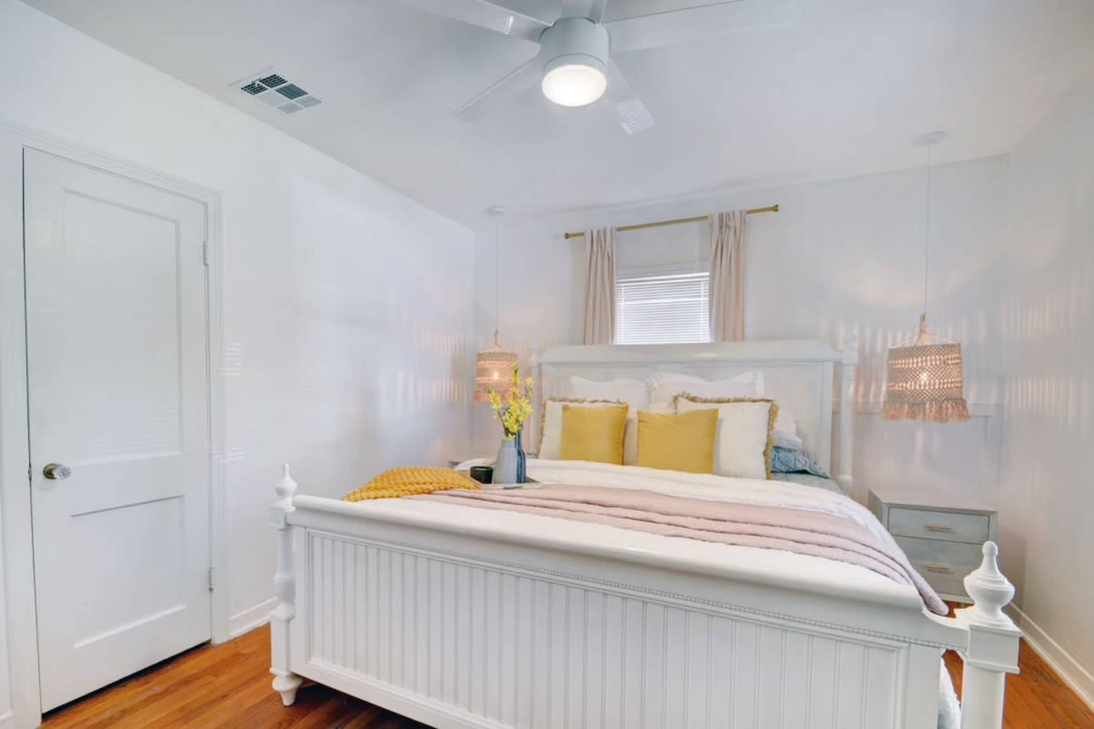 A brightly lit bedroom features a white bed with layered bedding and hanging pendant lights on either side.