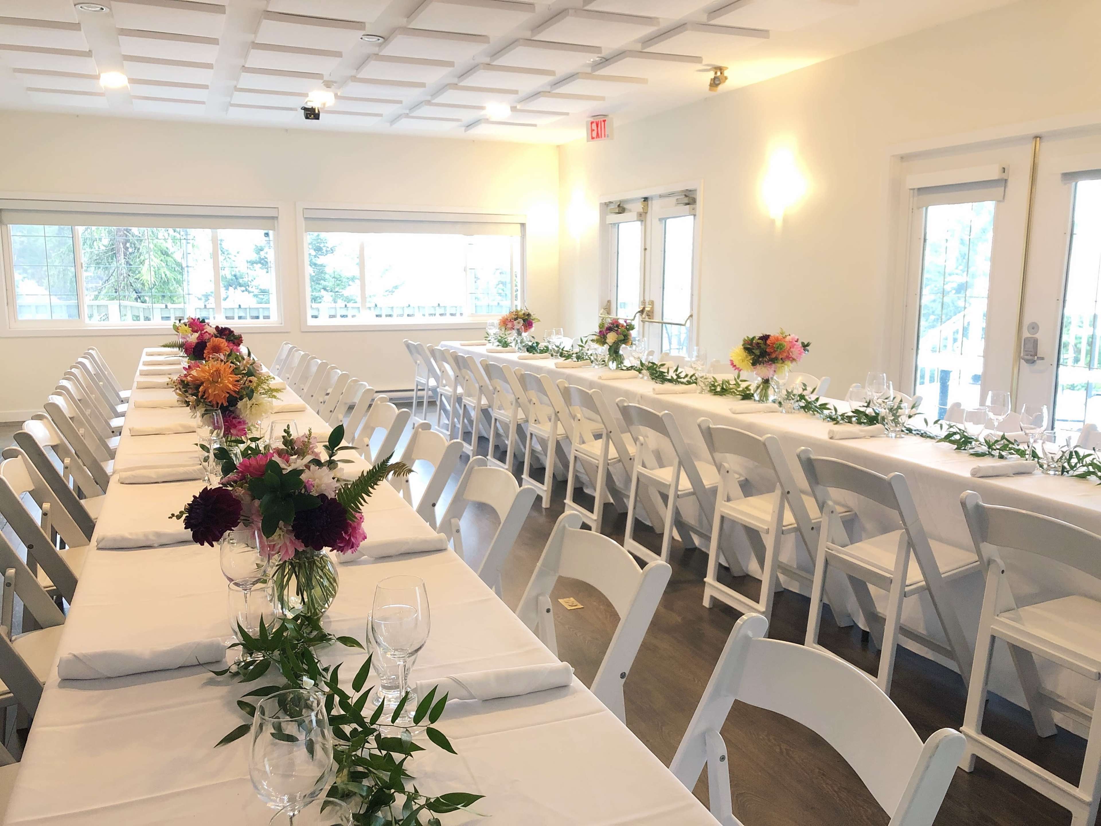 A spacious dining room features multiple long tables adorned with white tablecloths and centerpieces of colorful flowers.