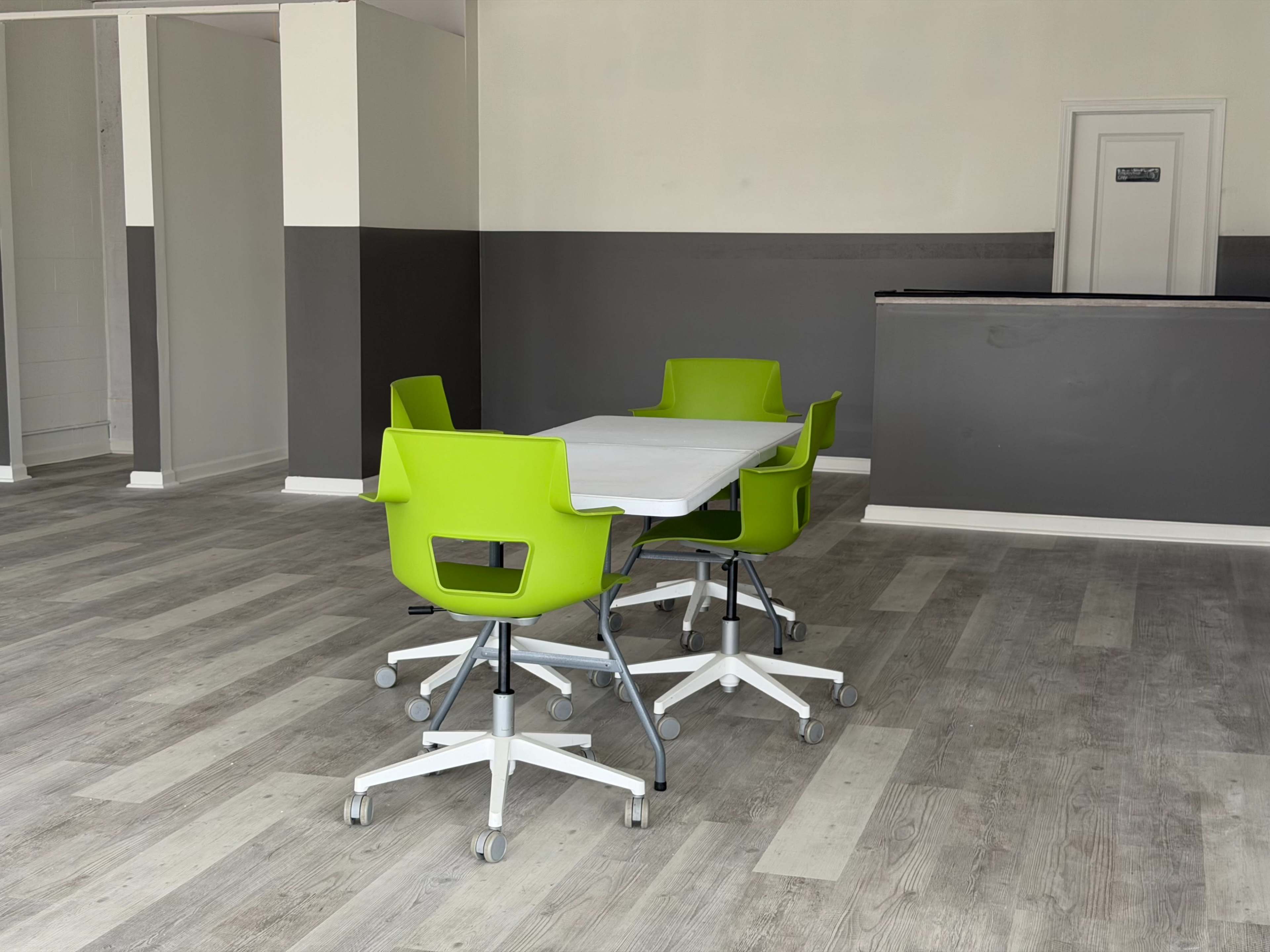A bright green table set with rolling chairs is positioned in a sparsely furnished room with gray walls and a reception area in the background.