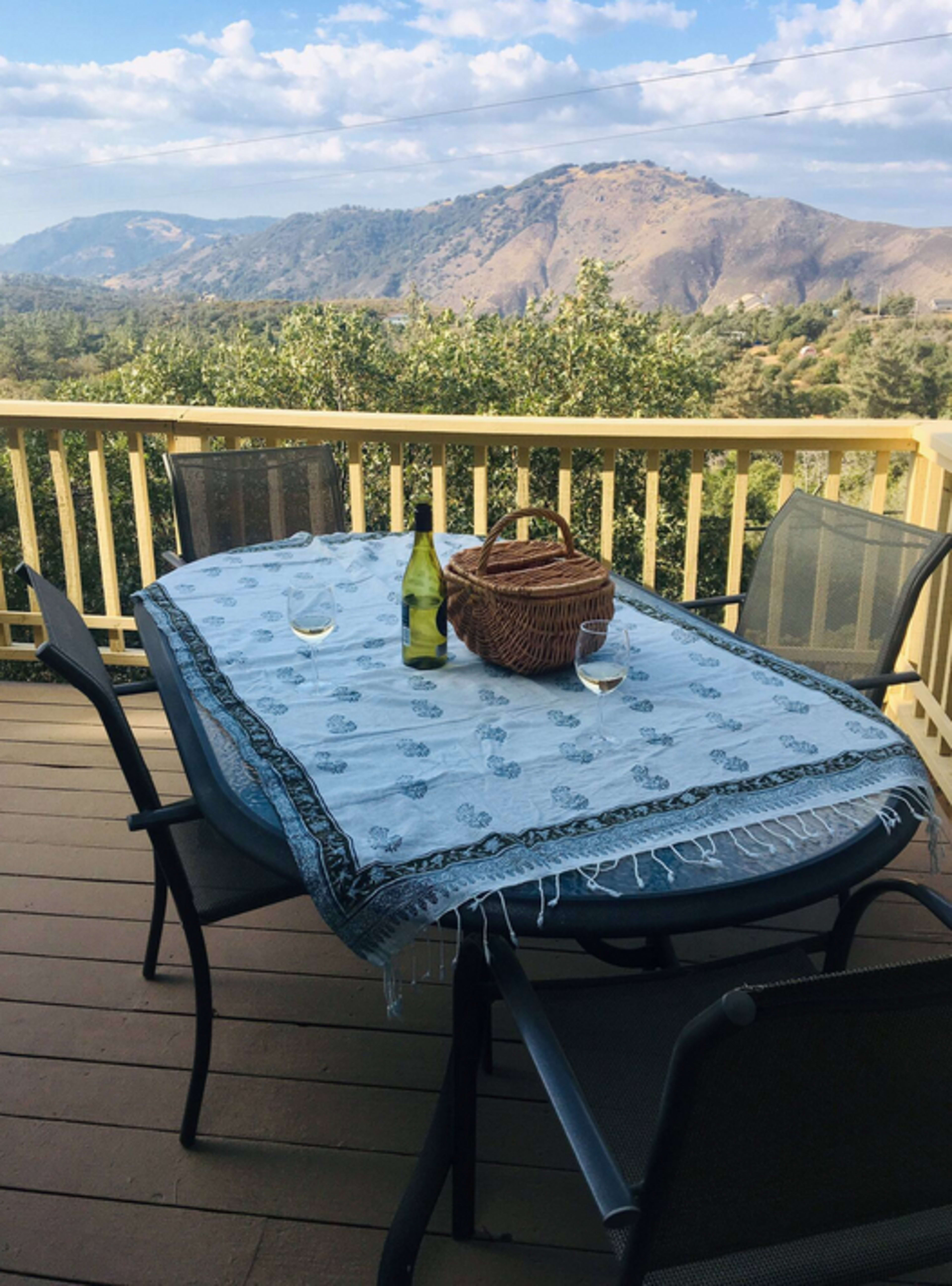 A dining table with a patterned tablecloth, two glasses, a wine bottle, and a wicker basket is set on a porch overlooking a mountainous landscape.