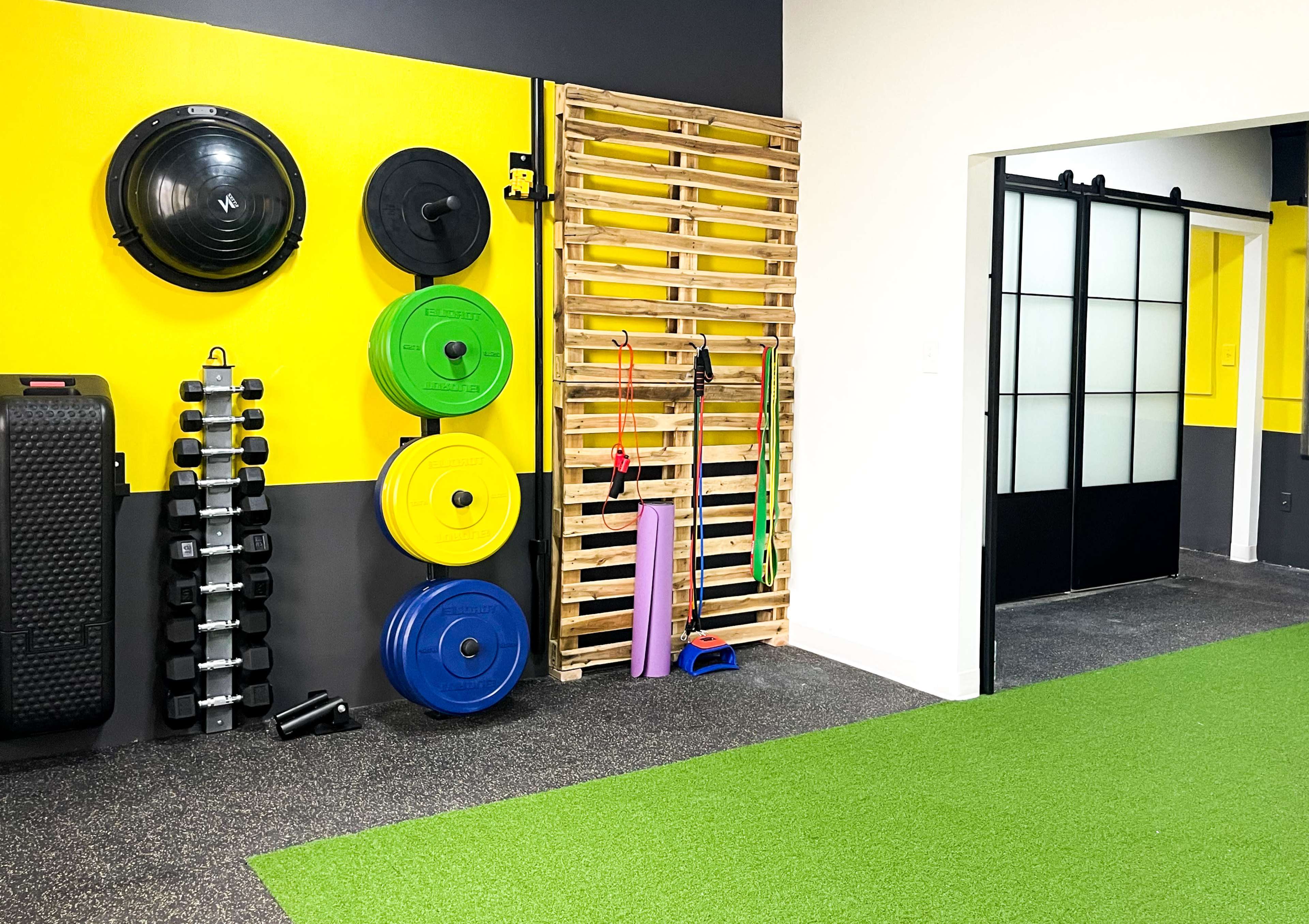A fitness space featuring colorful weight plates, dumbbells, resistance bands, and a wooden pallet for storage against a yellow and gray wall.