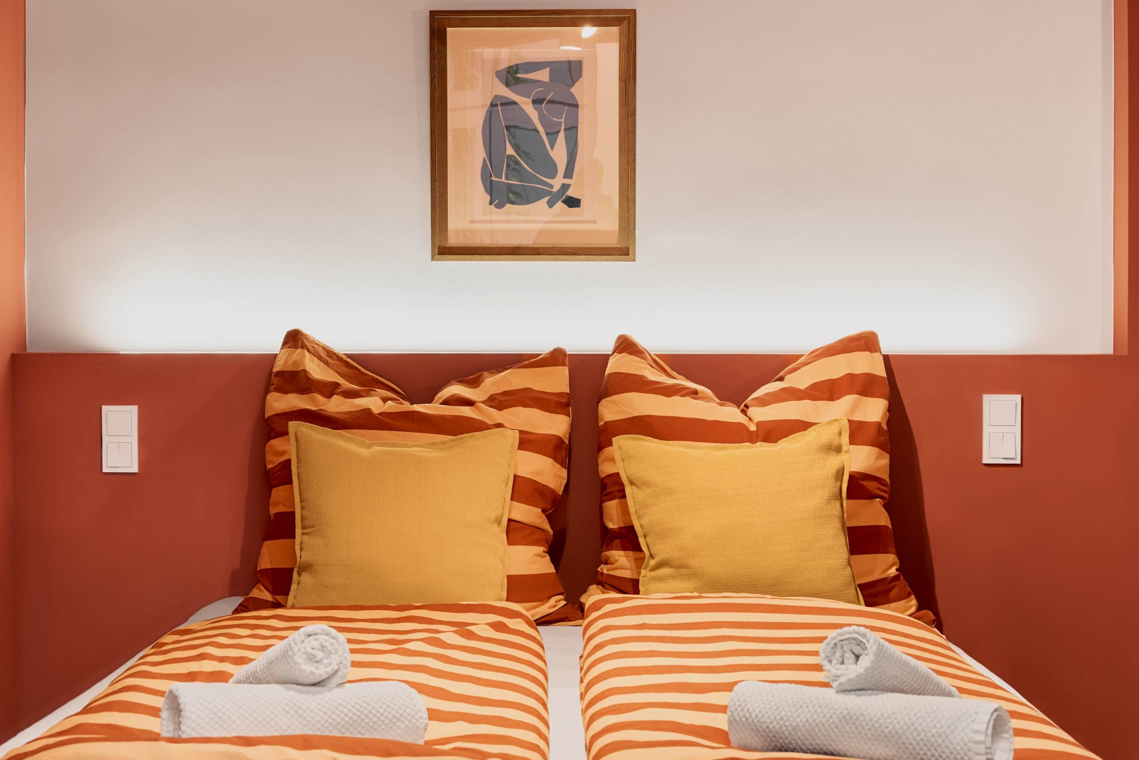 A bedroom features two twin beds with striped orange and white bedding, accented by large yellow pillows and a framed abstract artwork above the headboard.