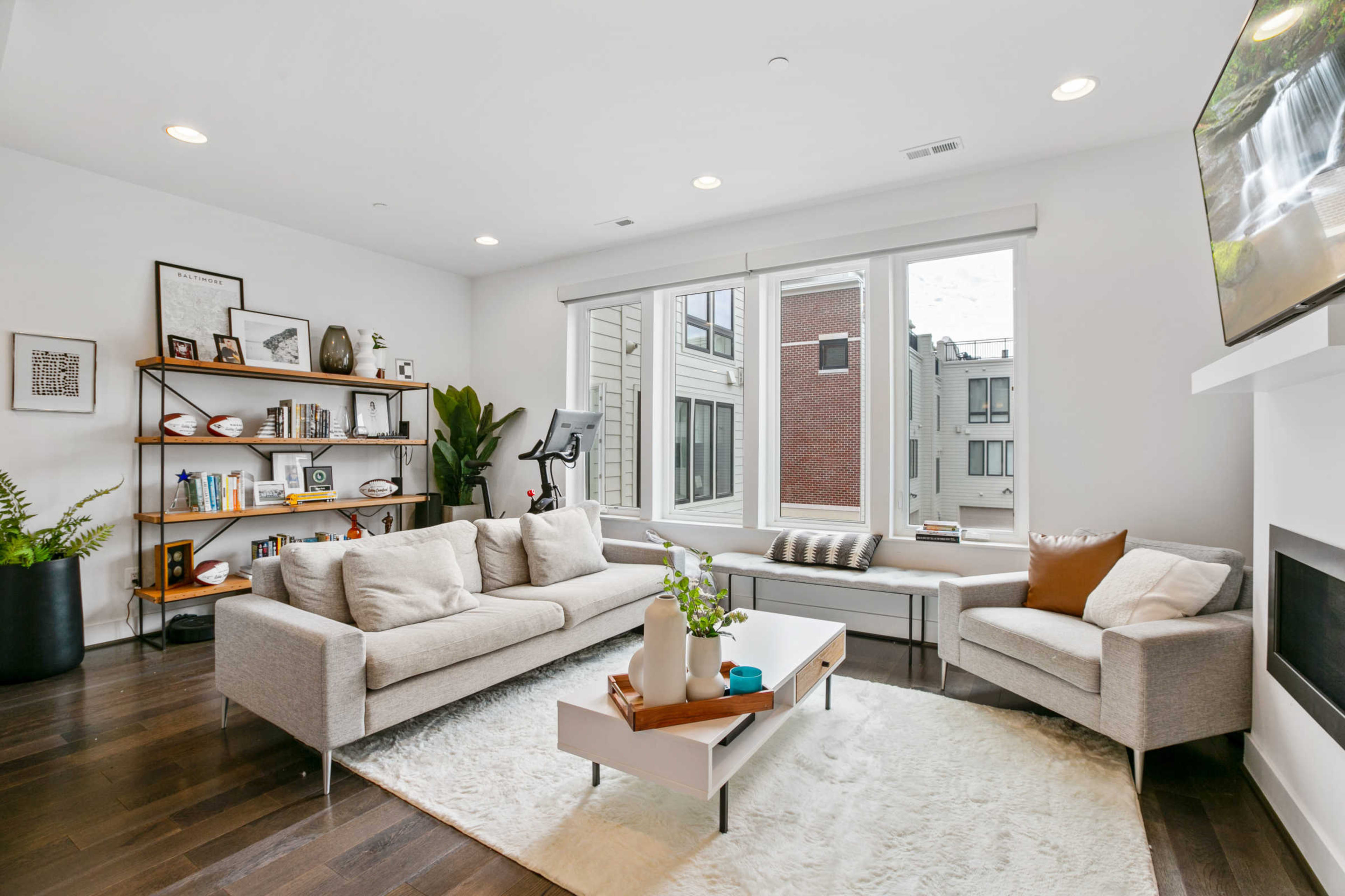 A modern living room with a light-colored sofa, a minimalist coffee table, large windows, and a bookshelf filled with decor and books.