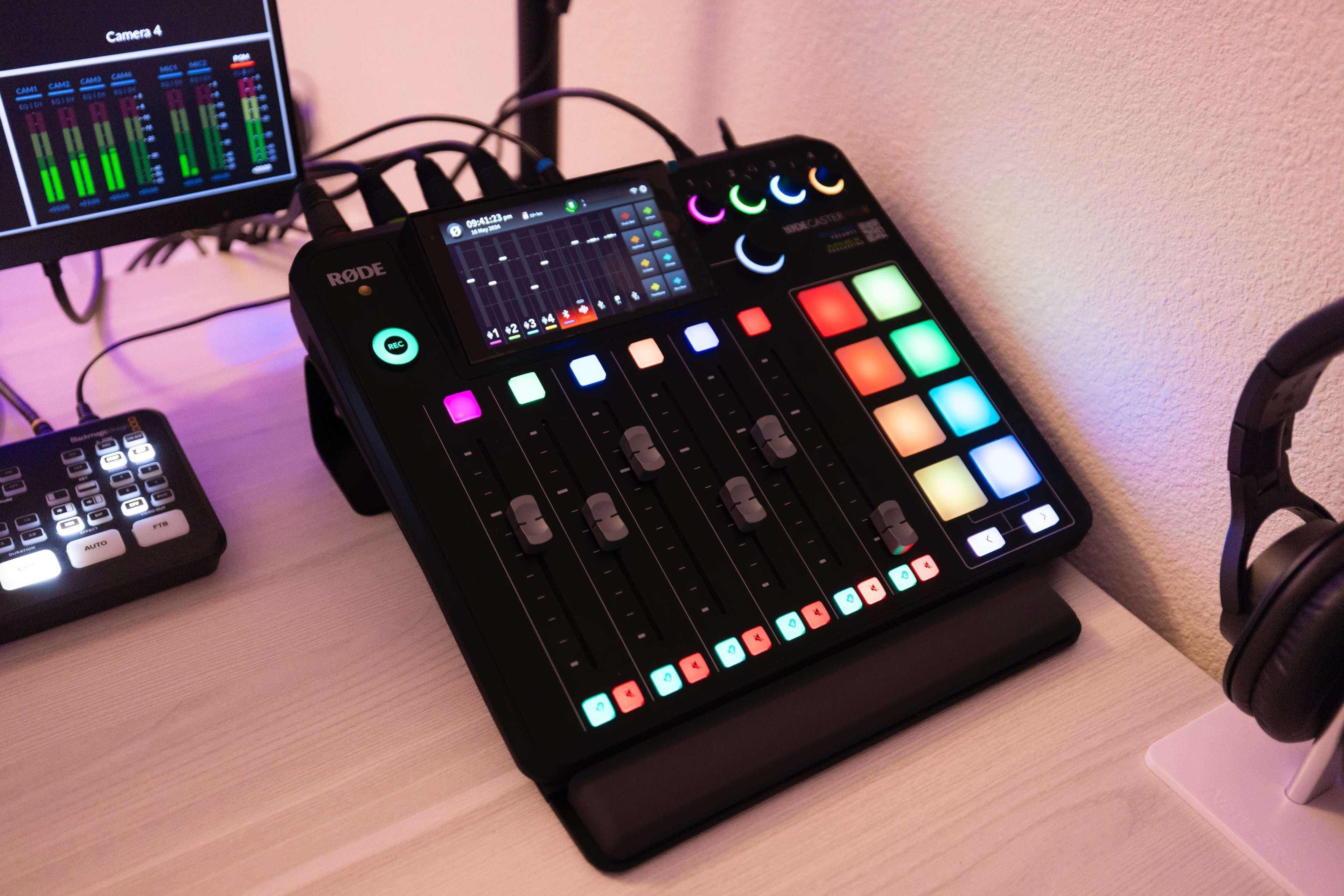 A Rode audio mixing console with multiple sliders, buttons, and a digital display, placed on a desk alongside a monitor.