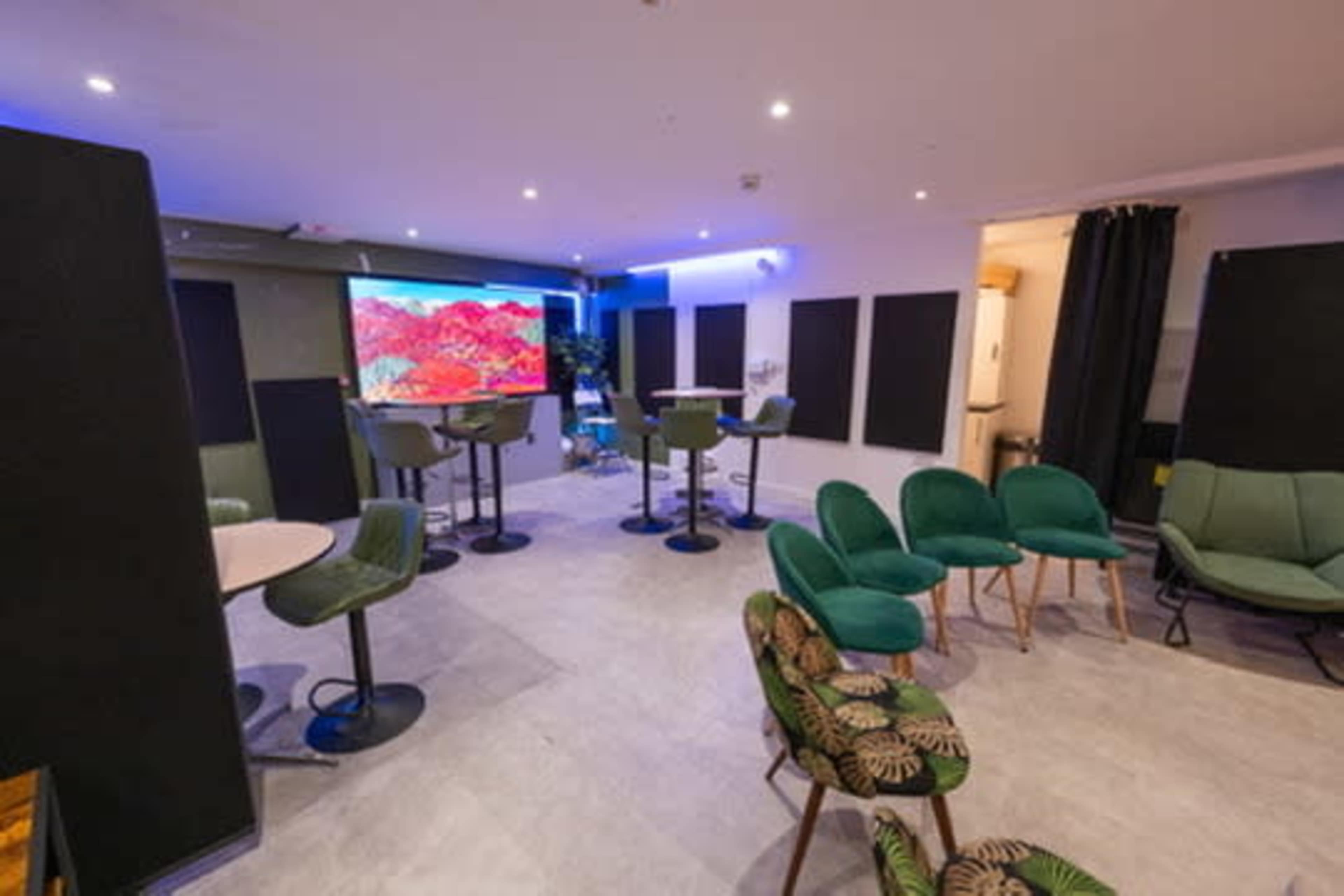 The image shows a modern lounge area with green chairs, bar stools, and a large screen displaying colorful visuals, set against a backdrop of dark panels and soft lighting.