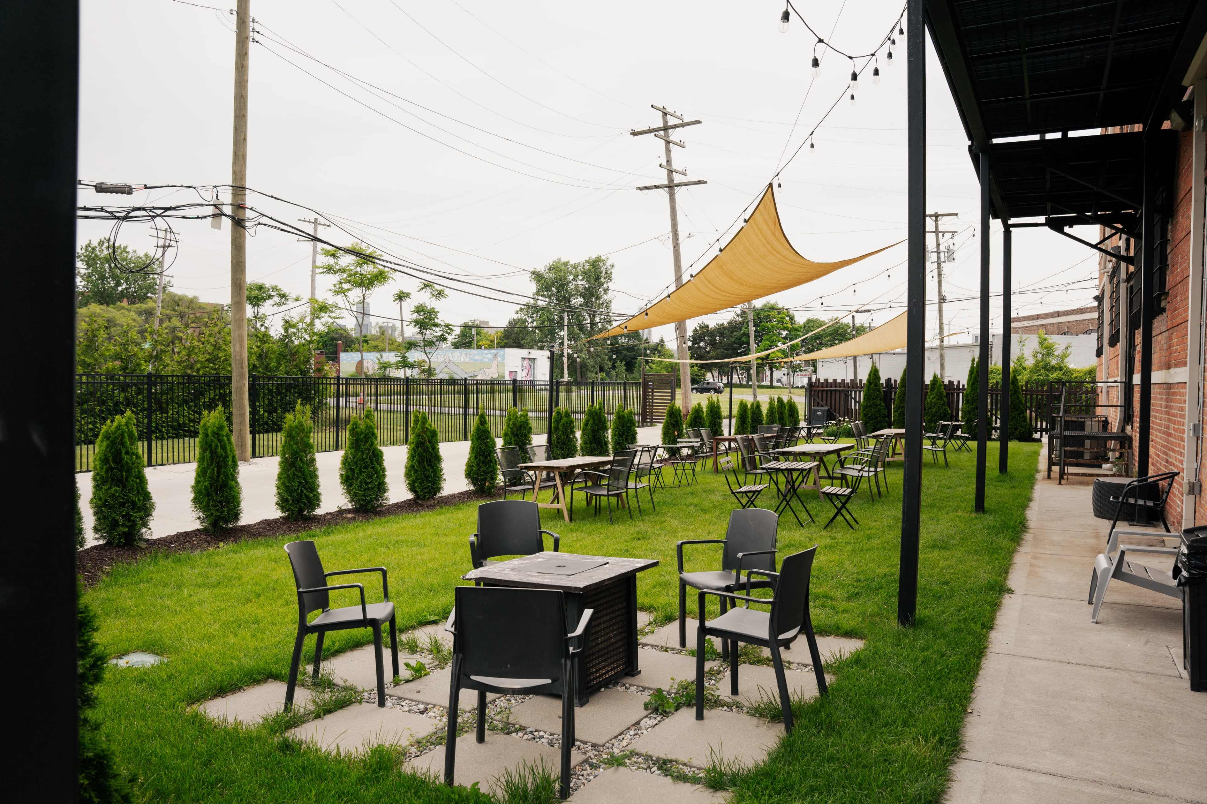 An outdoor seating area with metal chairs and tables surrounded by neatly arranged hedges and a large canopy providing shade.