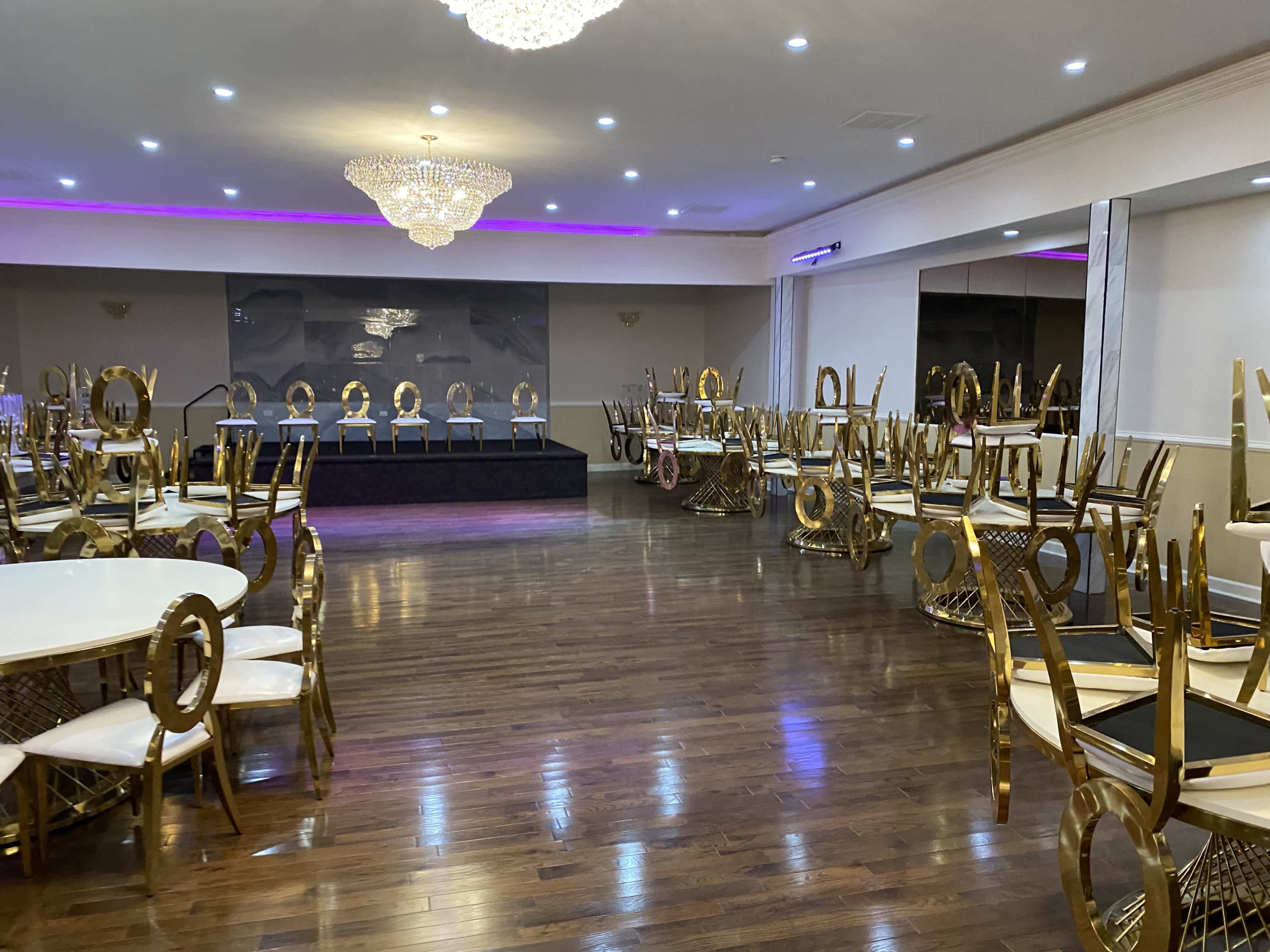 A banquet hall featuring empty circular tables with gold chairs stacked upside down, a stage at the back, and chandeliers overhead.