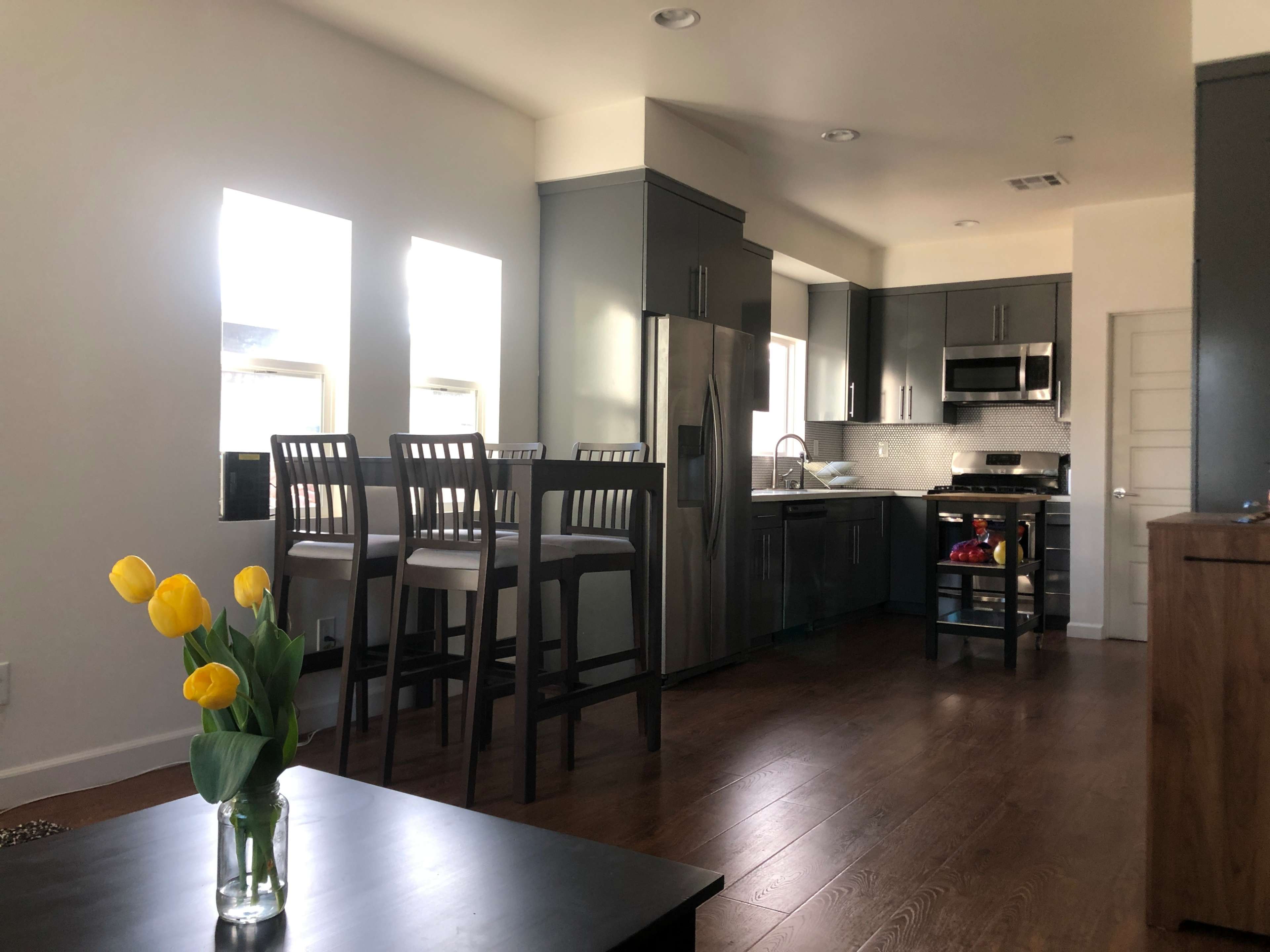 The image shows a modern kitchen with gray cabinetry, stainless steel appliances, and a dining area featuring tall chairs near the windows.