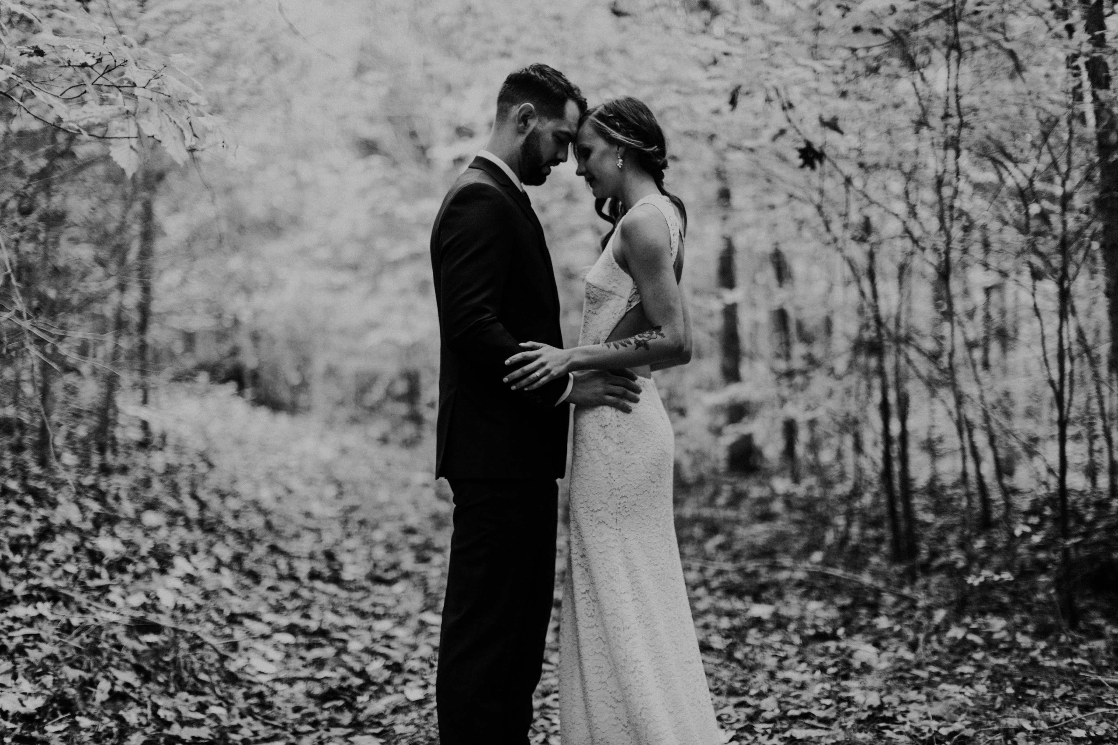 A couple stands close together in a forest, surrounded by trees and foliage.