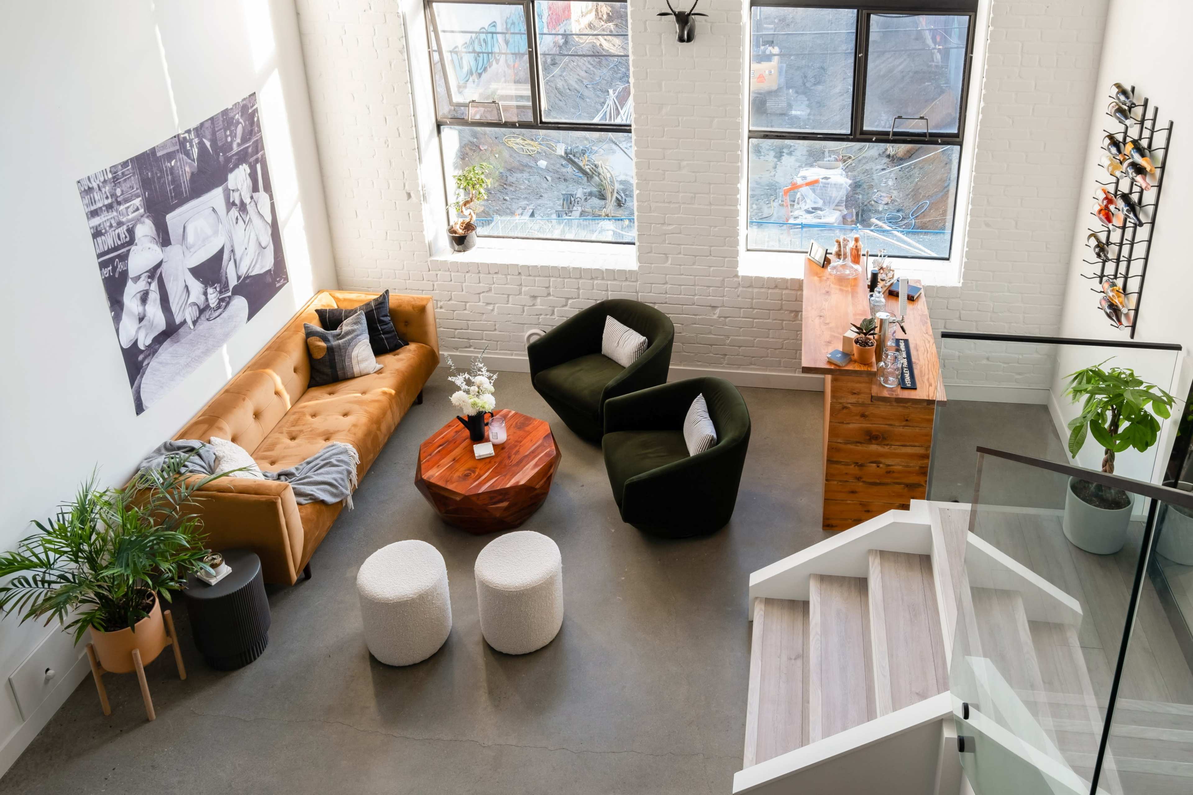 The scene depicts a modern living space with a tan leather sofa, two dark green armchairs, a wooden coffee table, and a bar area illuminated by large windows.