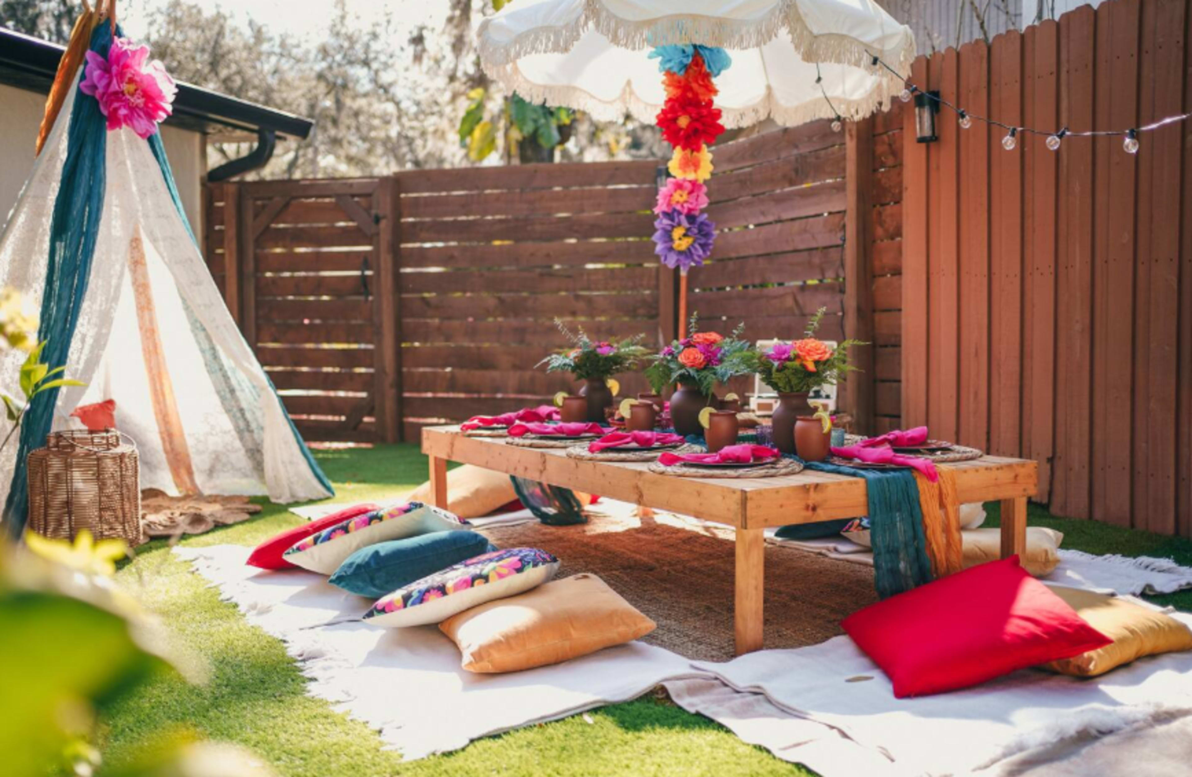 A vibrant outdoor setting features a wooden table adorned with colorful flowers and dishes, surrounded by plush cushions and a decorative umbrella.
