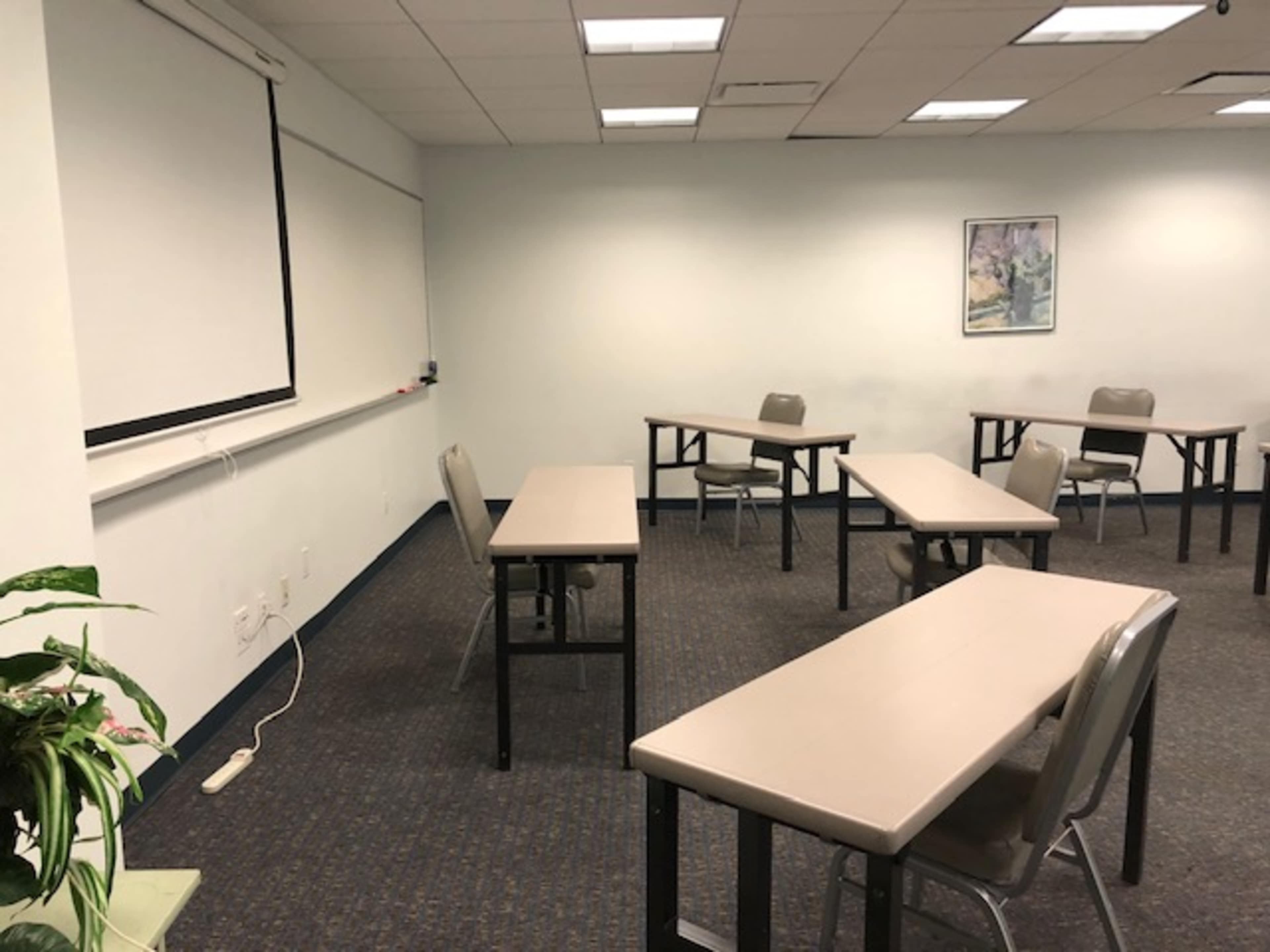 A classroom with several tables and chairs arranged for seating, featuring a blank projector screen on one wall.