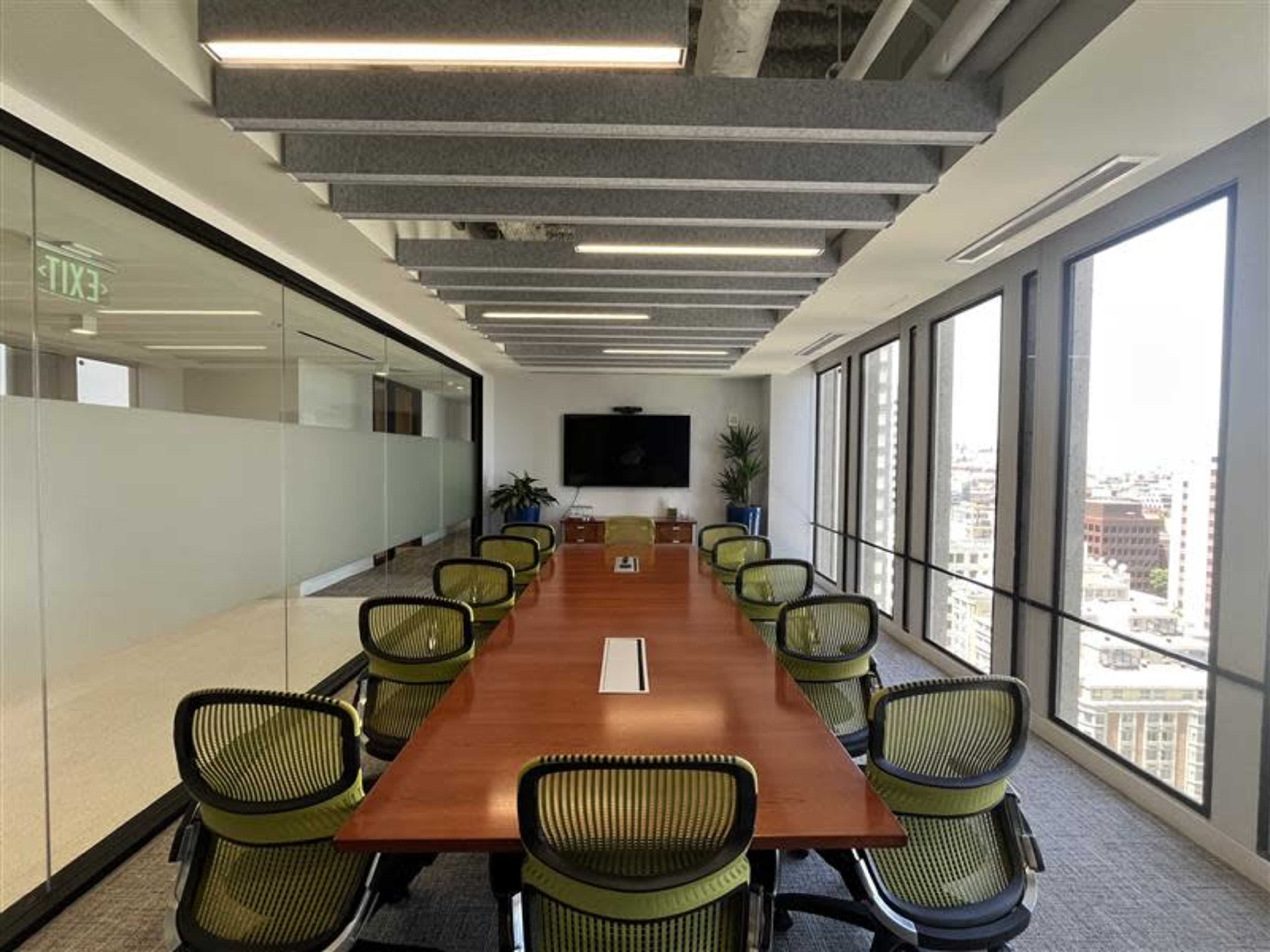 The image shows a modern conference room with a long wooden table surrounded by green office chairs, large windows providing a city view, and a TV mounted on the wall.