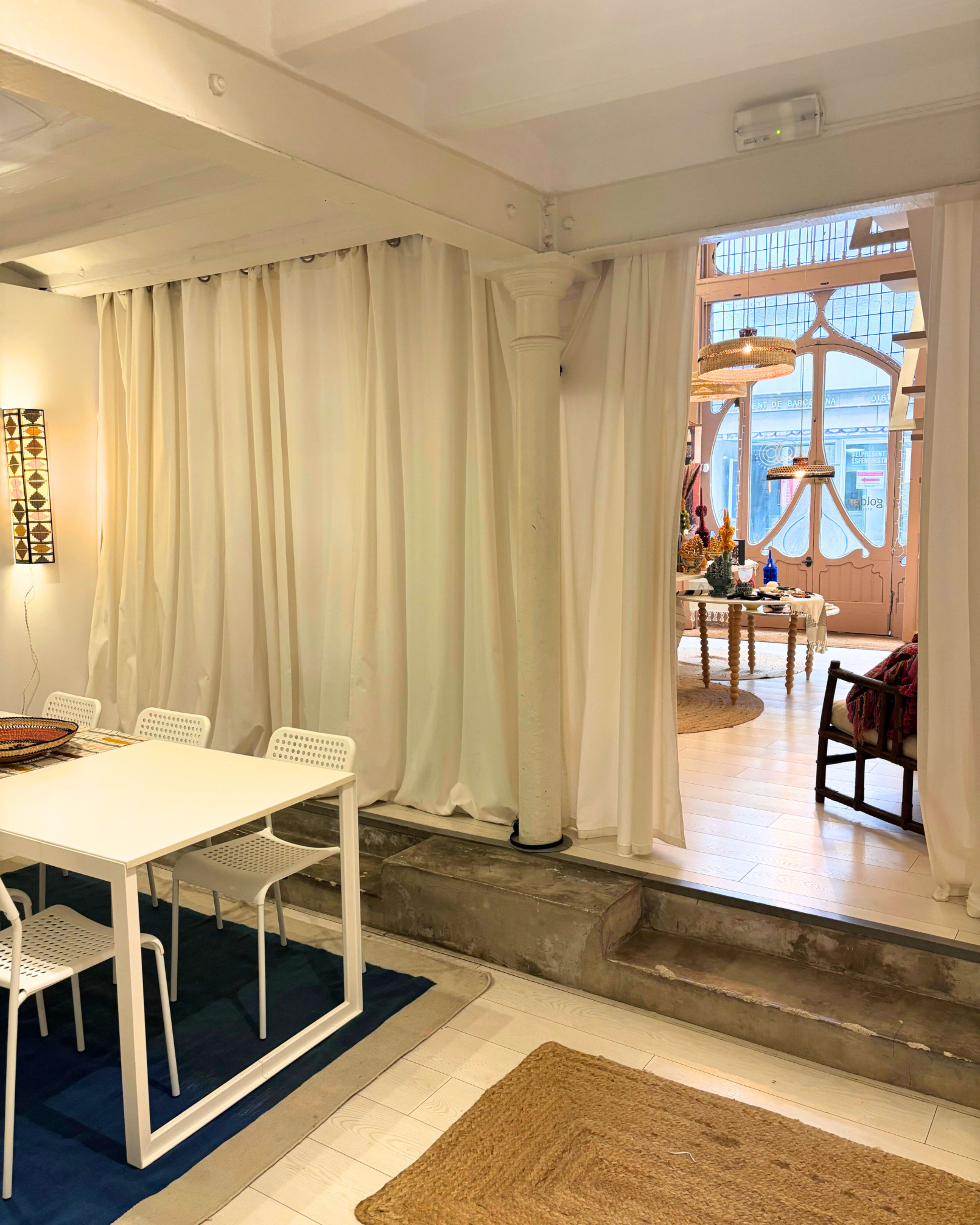 A dining area with a white table and four chairs is separated from a brightly lit room by white curtains and features a woven rug on the floor.