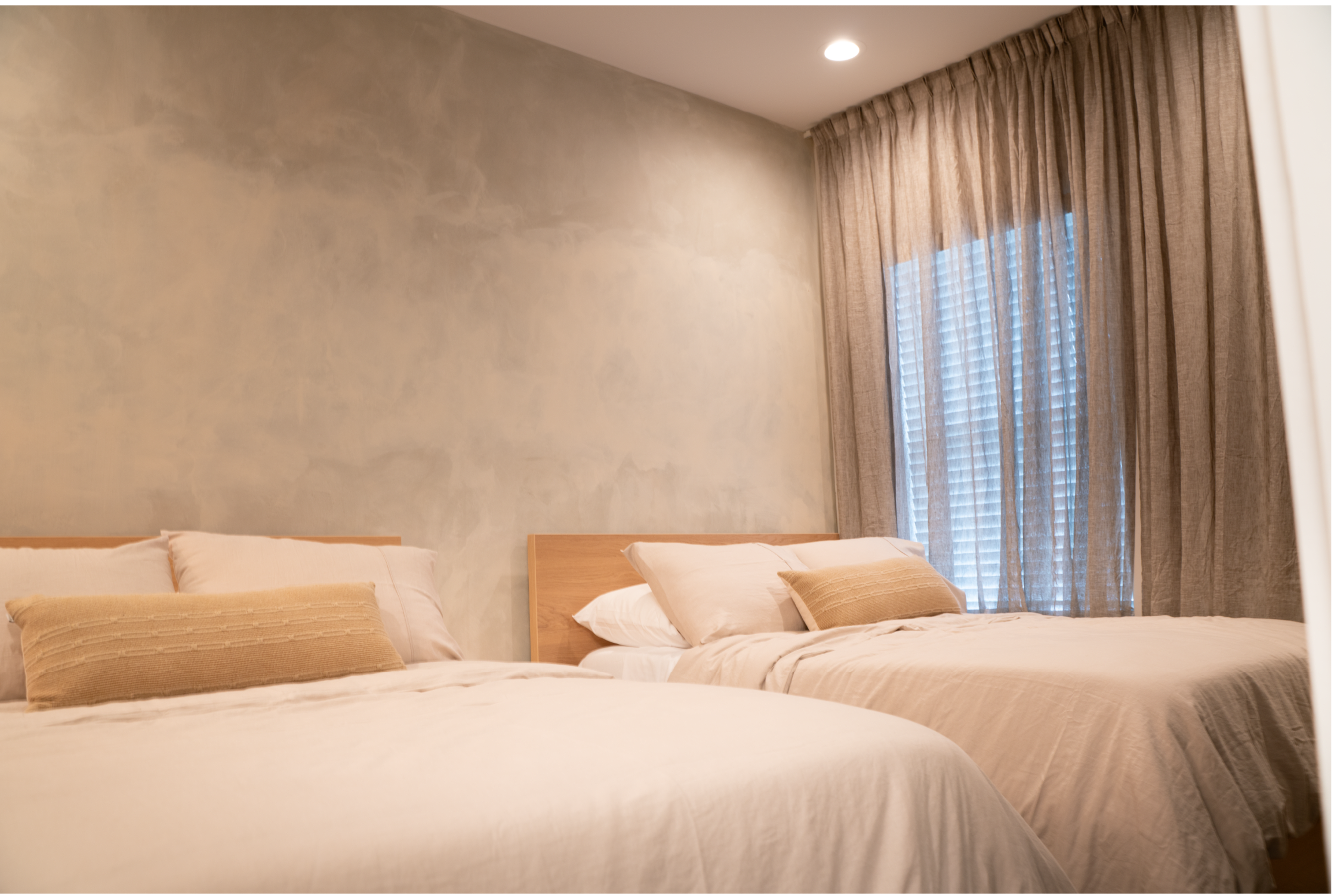 The image shows a well-lit room featuring two beds with light-colored bedding and a large window covered with beige curtains.
