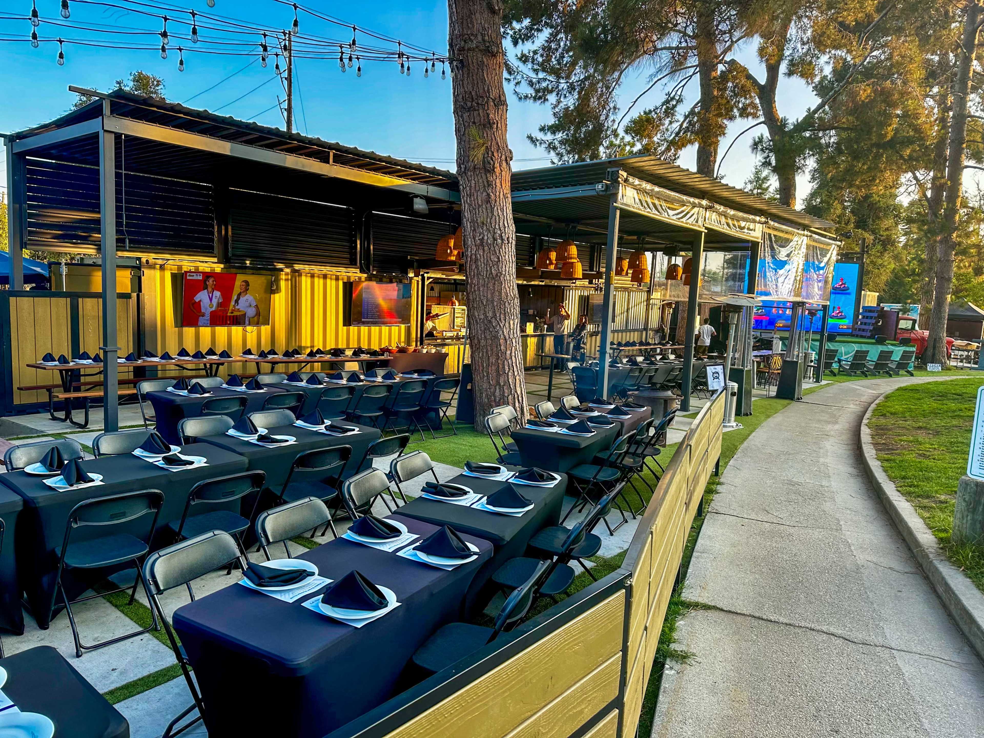 The image shows an outdoor dining area with rows of black tables and folding chairs next to a walkway, surrounded by trees and string lights.