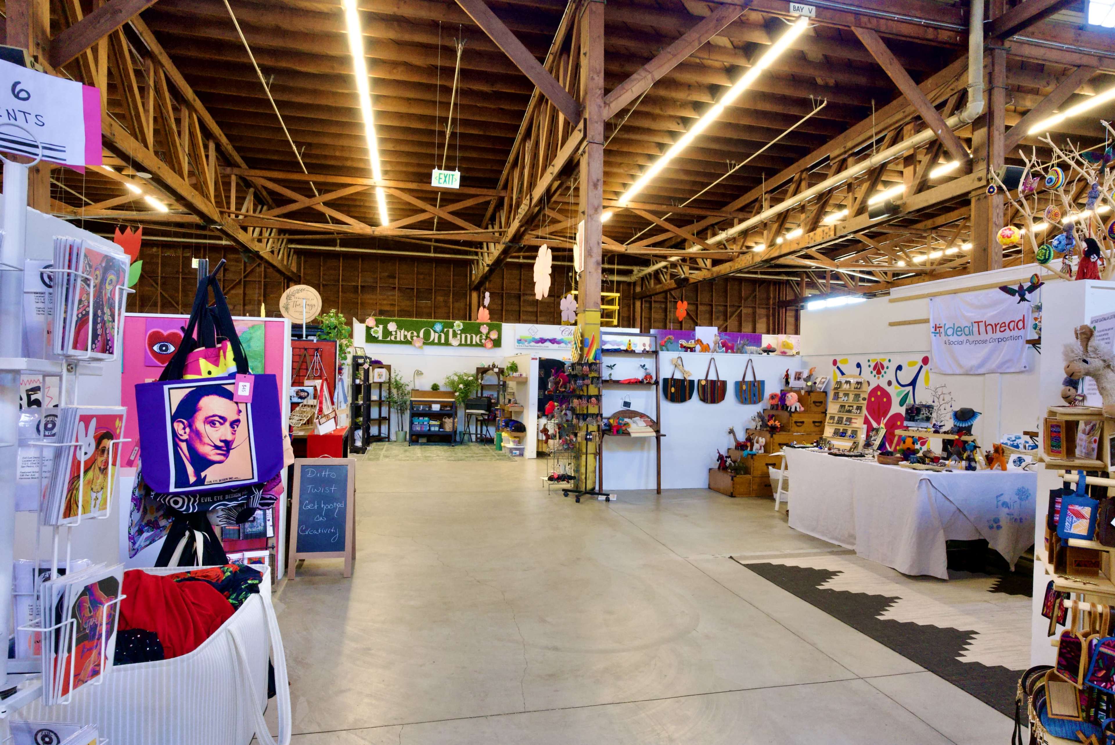The image shows a spacious indoor market with various colorful stalls and displays featuring art, crafts, and merchandise.