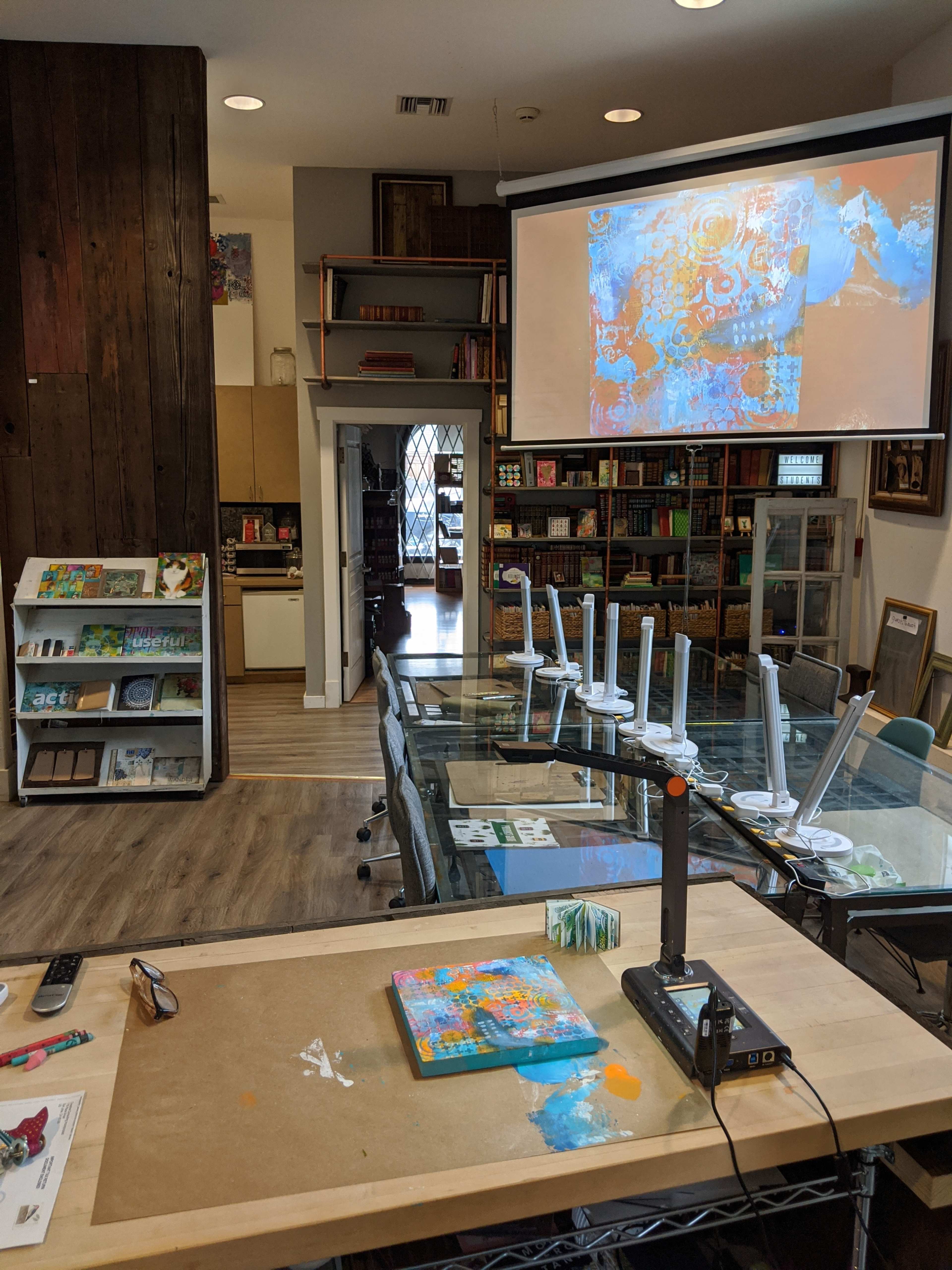 A bright art studio with a large table in the foreground, a projector displaying artwork on the wall, and shelves filled with art supplies in the background.