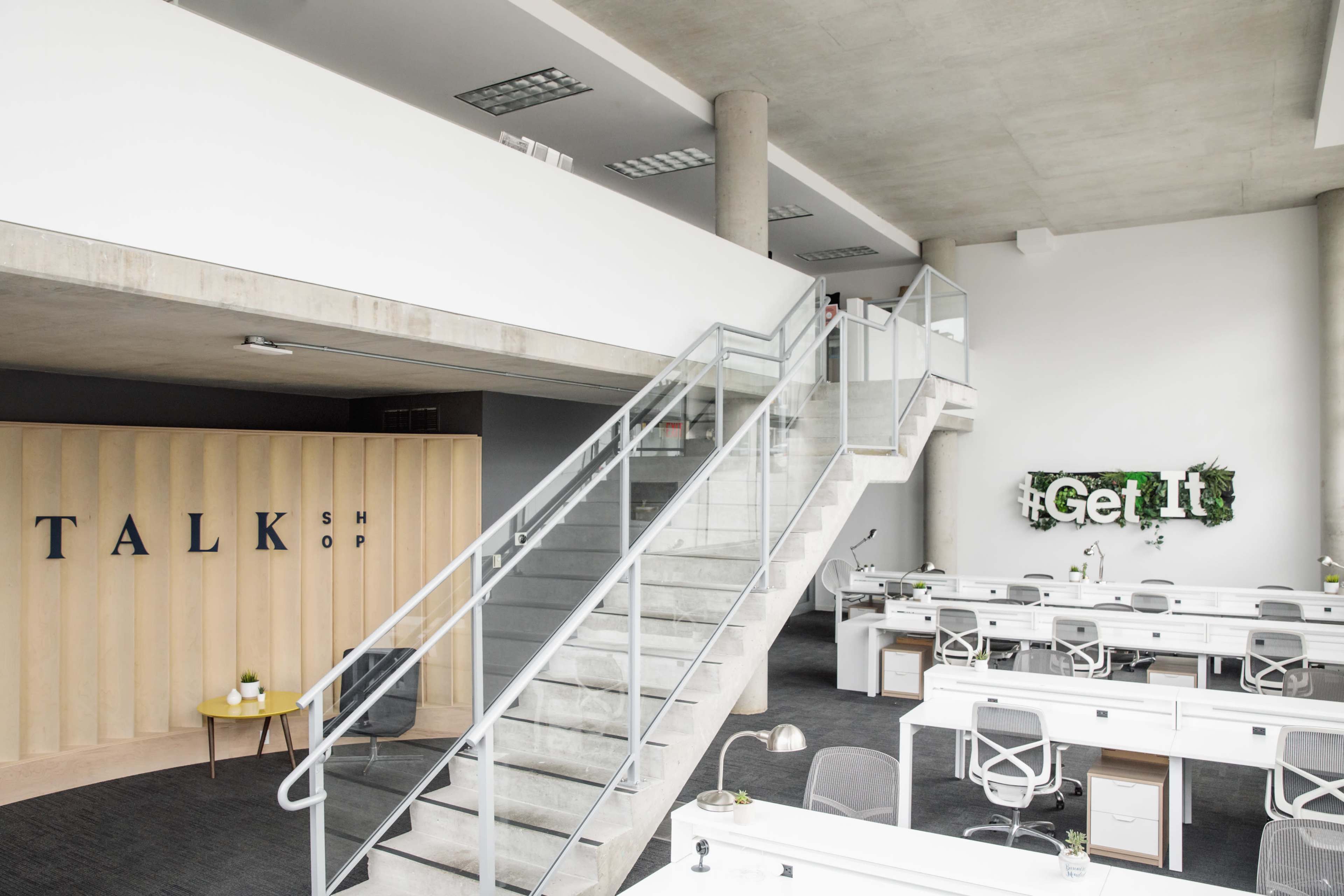 A modern office space with a staircase, a "TALK" sign on a wooden wall, and several workstations equipped with desks and plants.