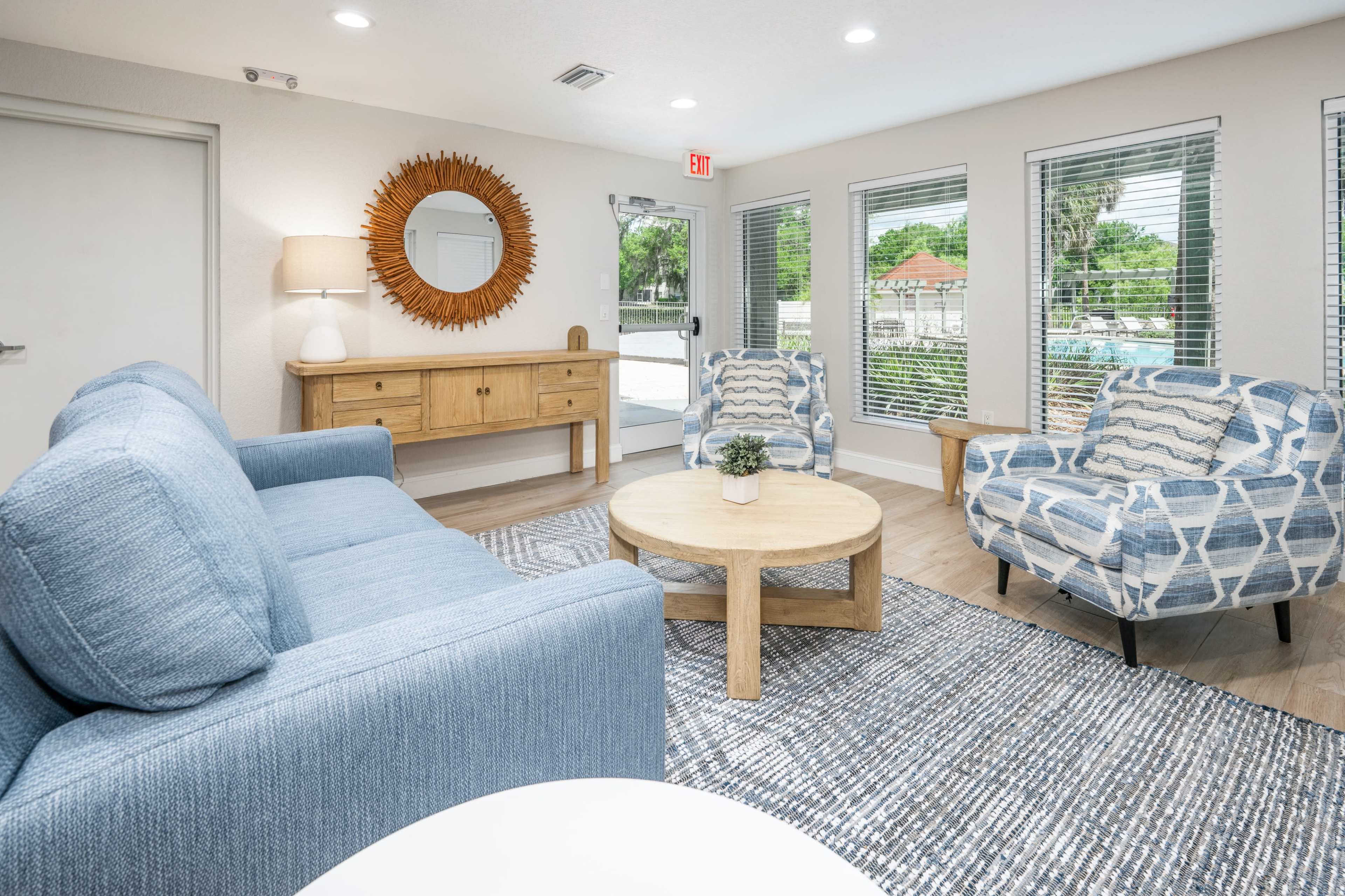 The image shows a modern lounge area with a blue sofa, two patterned armchairs, a round coffee table, and a sideboard with a mirror, all set around large windows that allow natural light to enter.