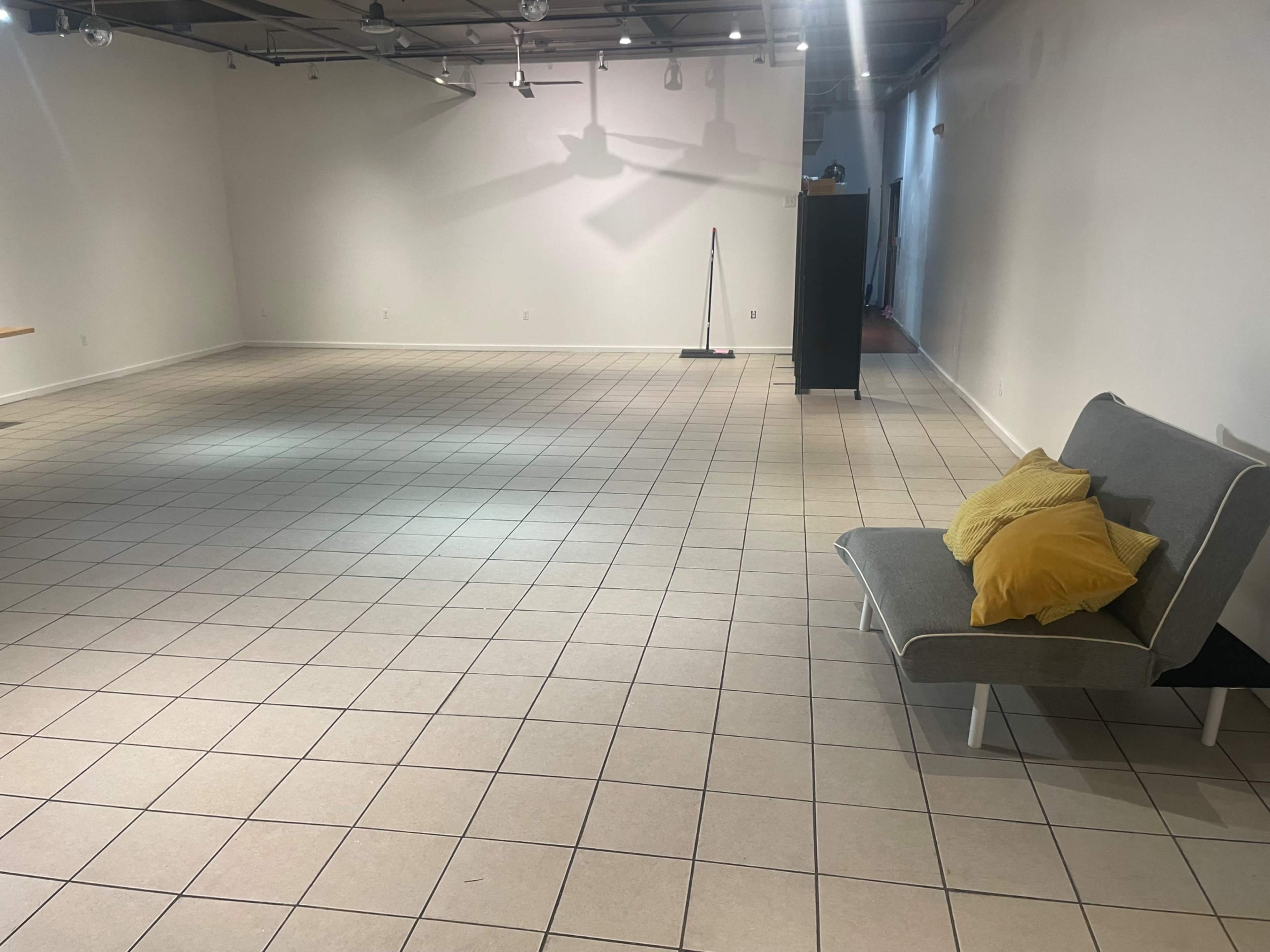A spacious, empty room with tiled flooring, a gray couch with yellow cushions, and minimal furniture.