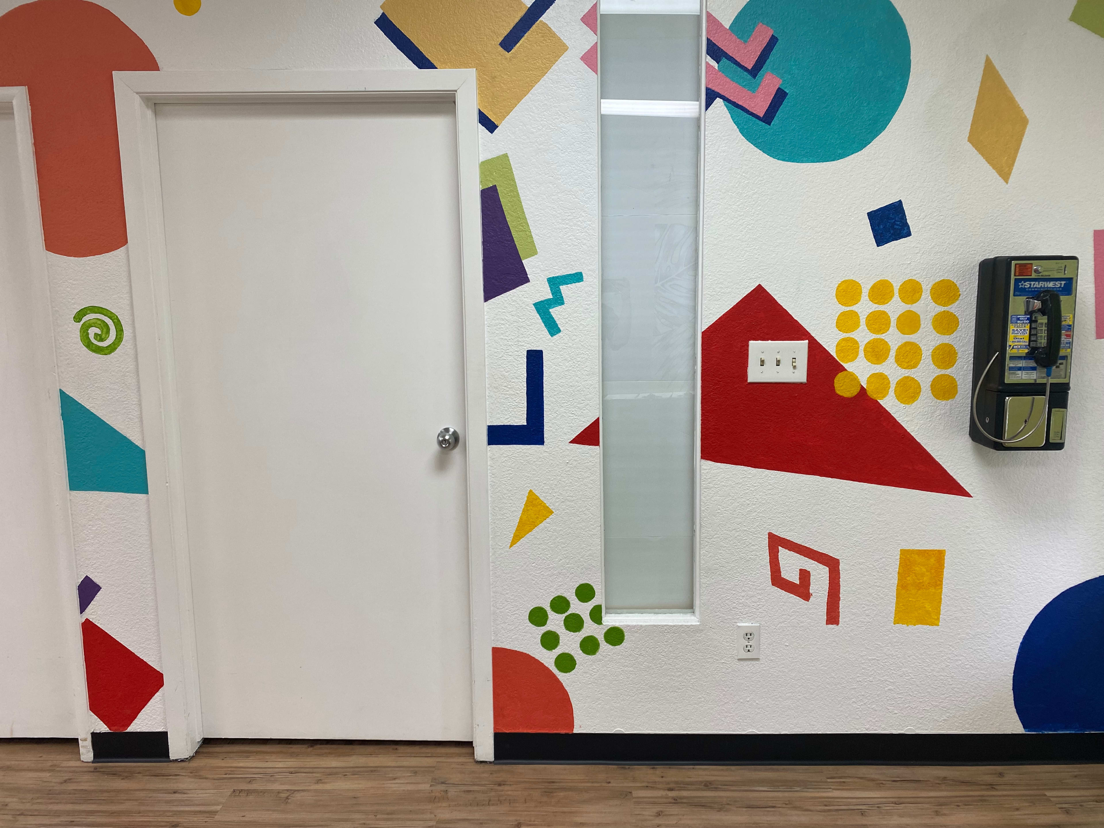 The image shows a brightly painted wall with geometric shapes in various colors next to a white door and a payphone.