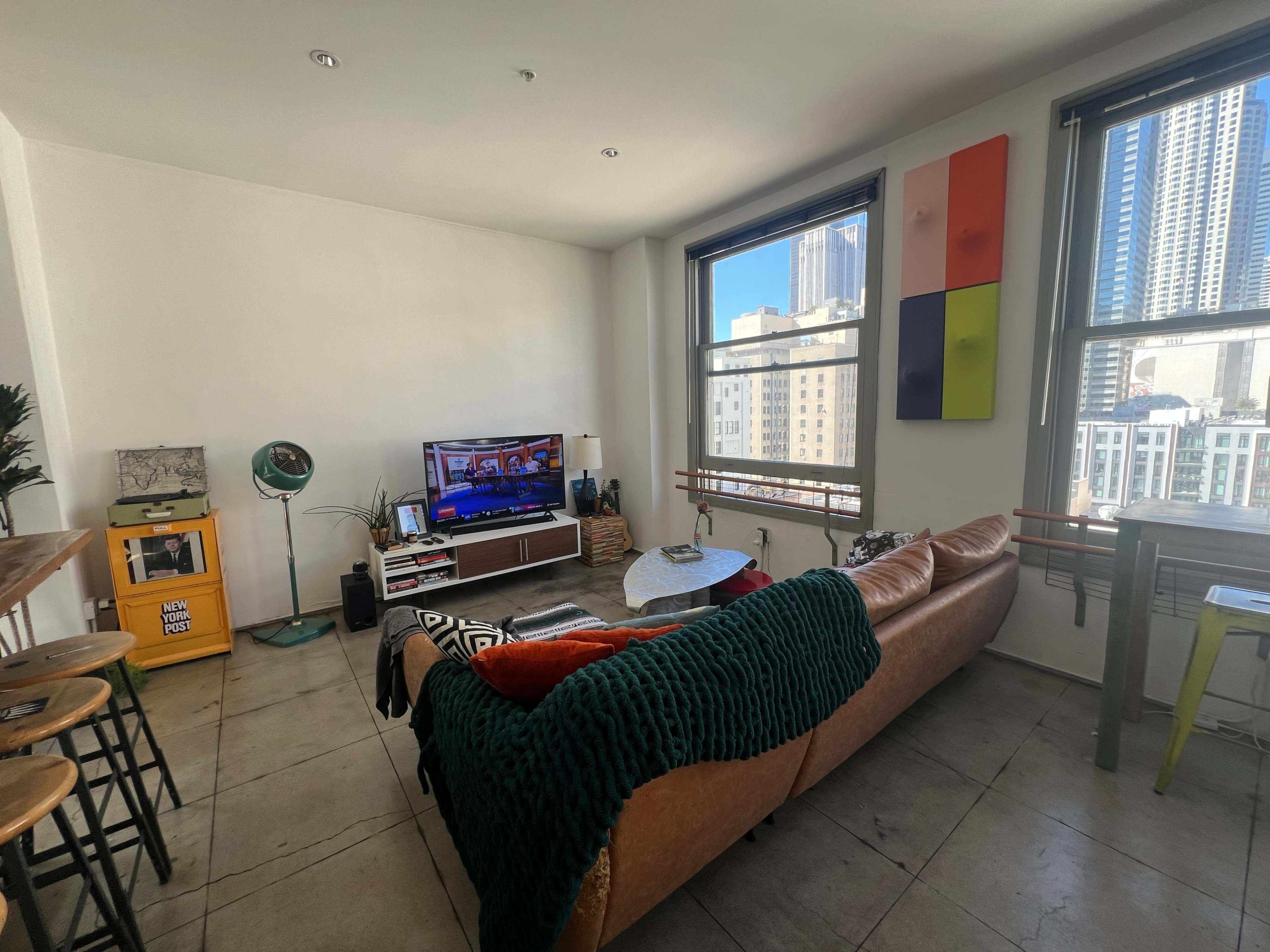 A living room featuring a brown leather sofa, a television on a media console, and large windows with city views.